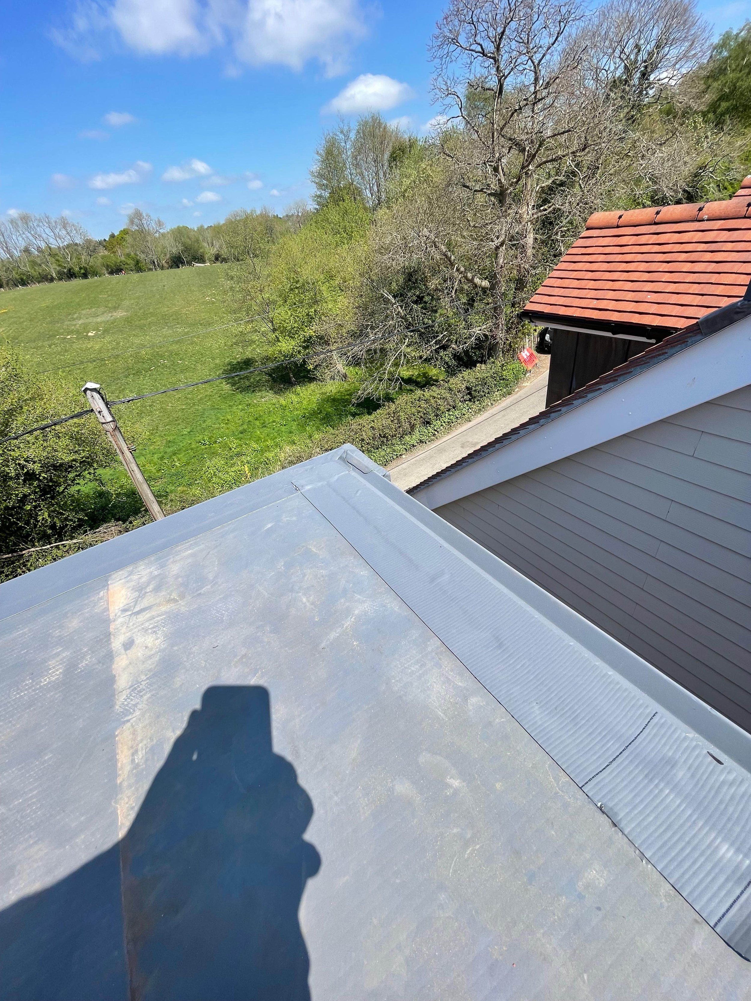 View from a rooftop showing a grassy hill, trees, a partly cloudy blue sky, and neighboring rooftops with red and gray shingles.