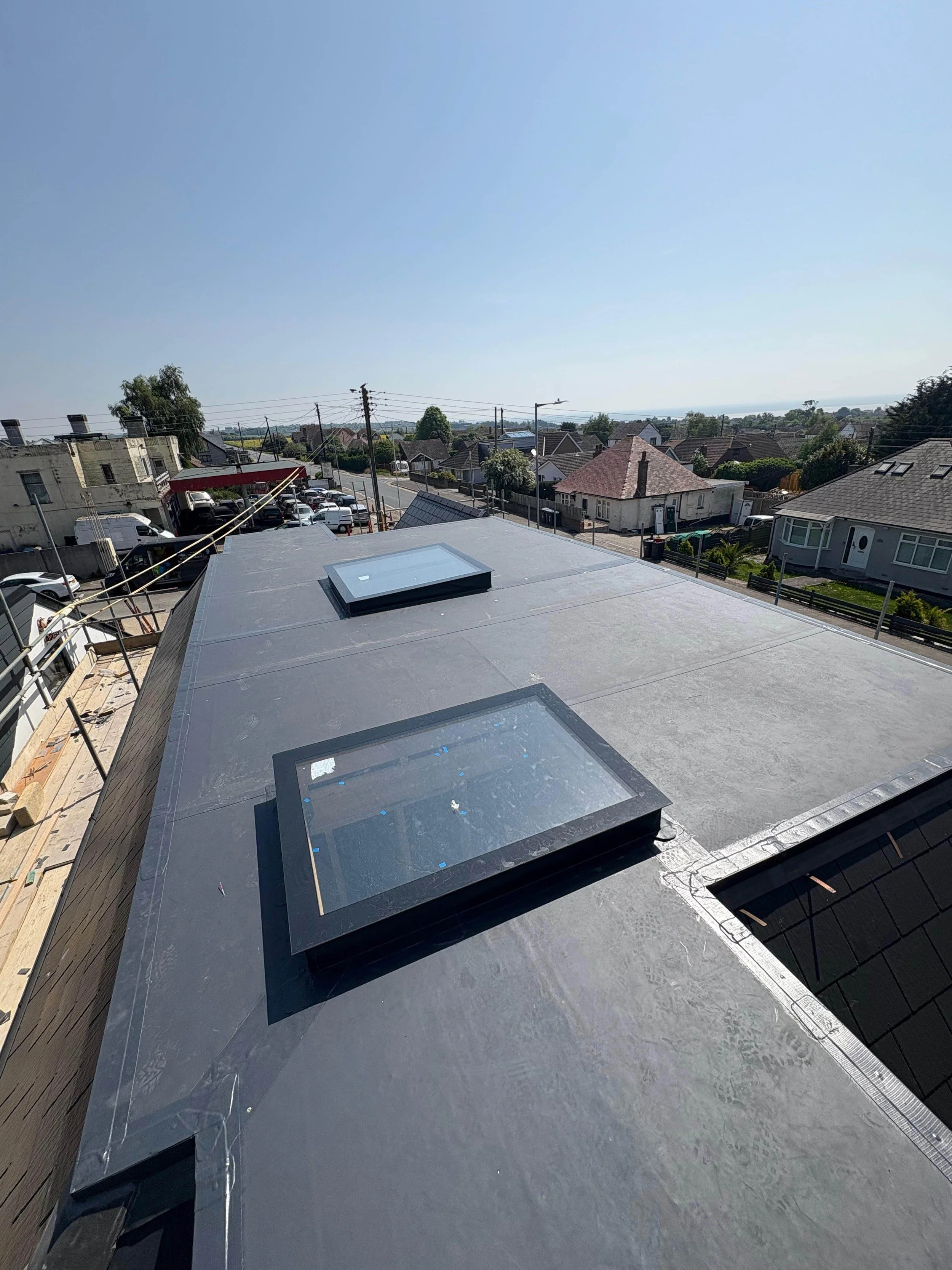View of a flat roof with two skylights, surrounded by residential houses, power lines, and a clear blue sky.