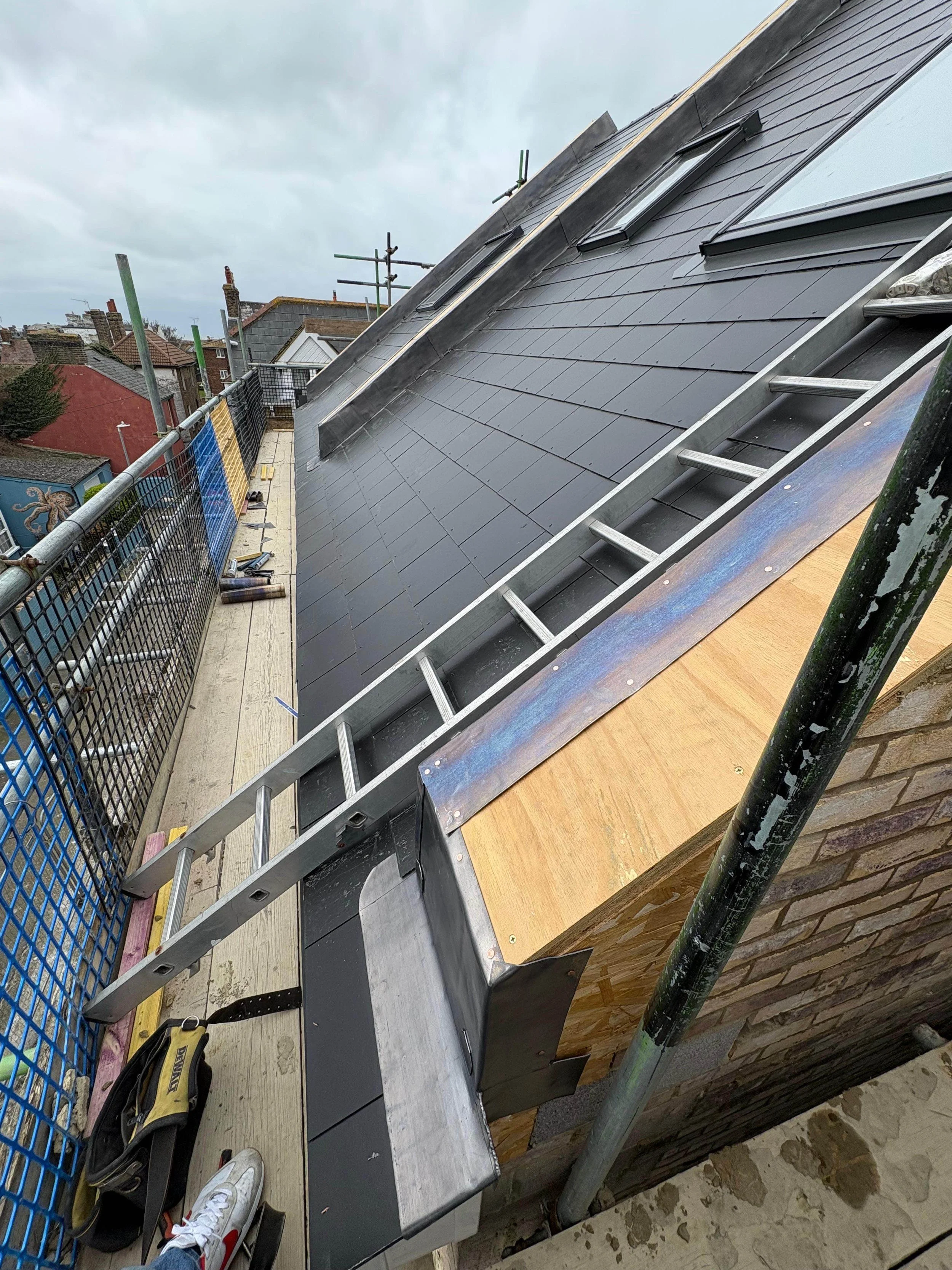 Construction site on a rooftop with a ladder, metal roofing materials, and scaffolding, overlooking neighboring buildings with cloudy sky.