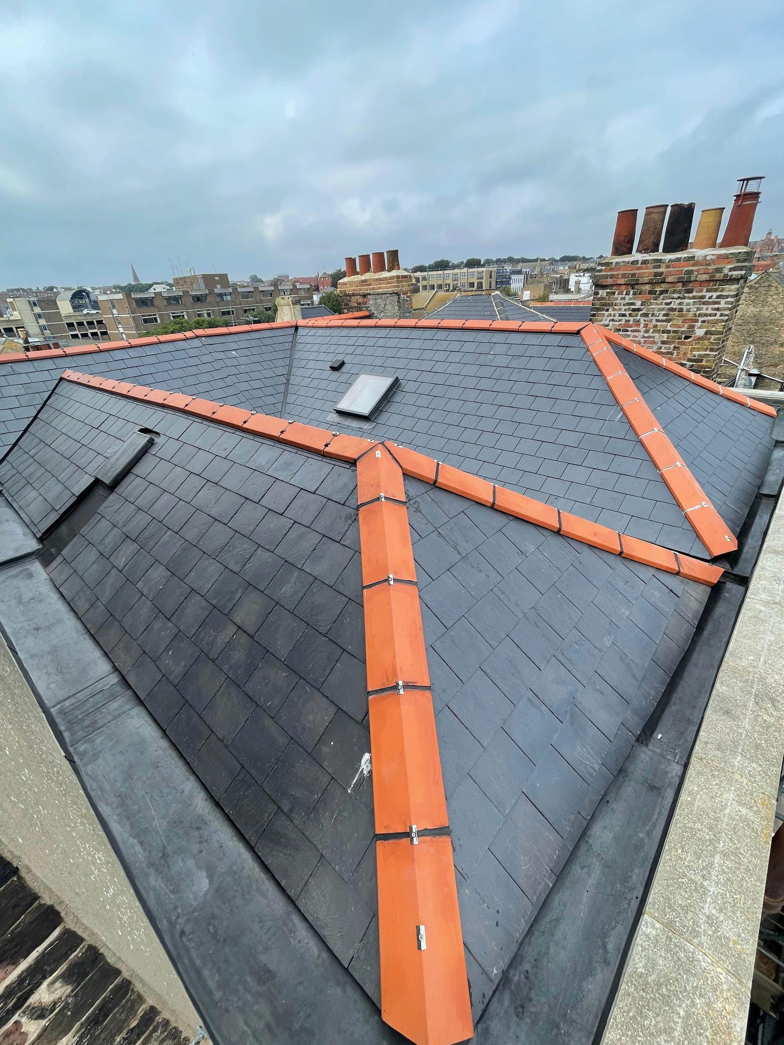 View of a rooftop with black tiles and orange ridge tiles, with neighboring rooftops and a cloudy sky in the background.