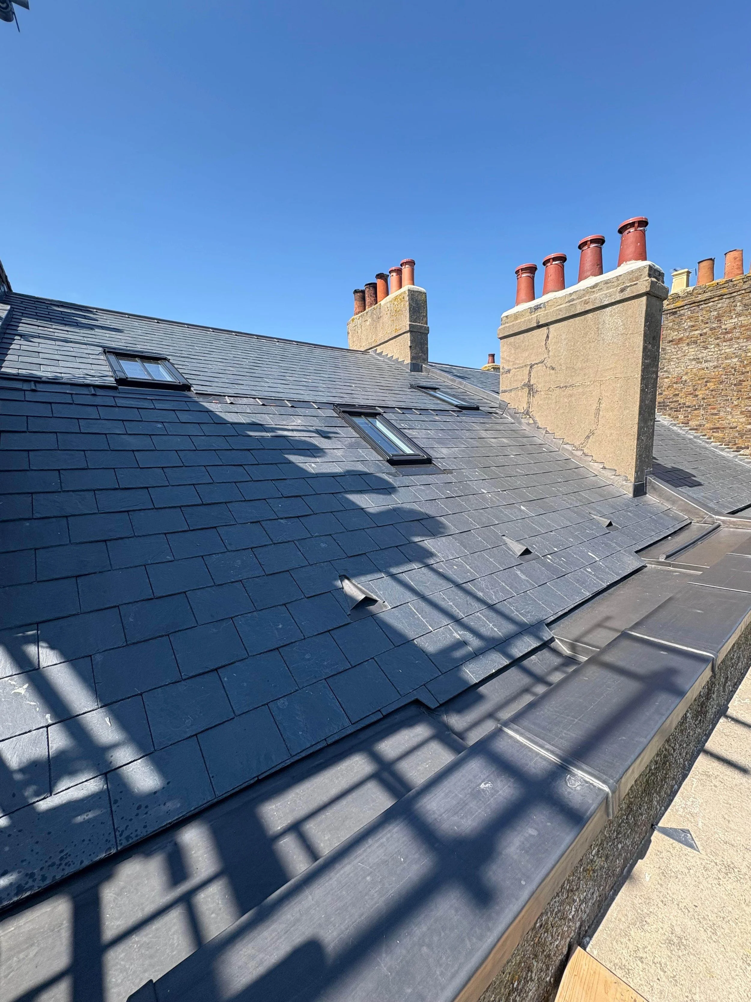 Rooftop with black slate tiles, two skylights, multiple red brick chimneys, and a clear blue sky.