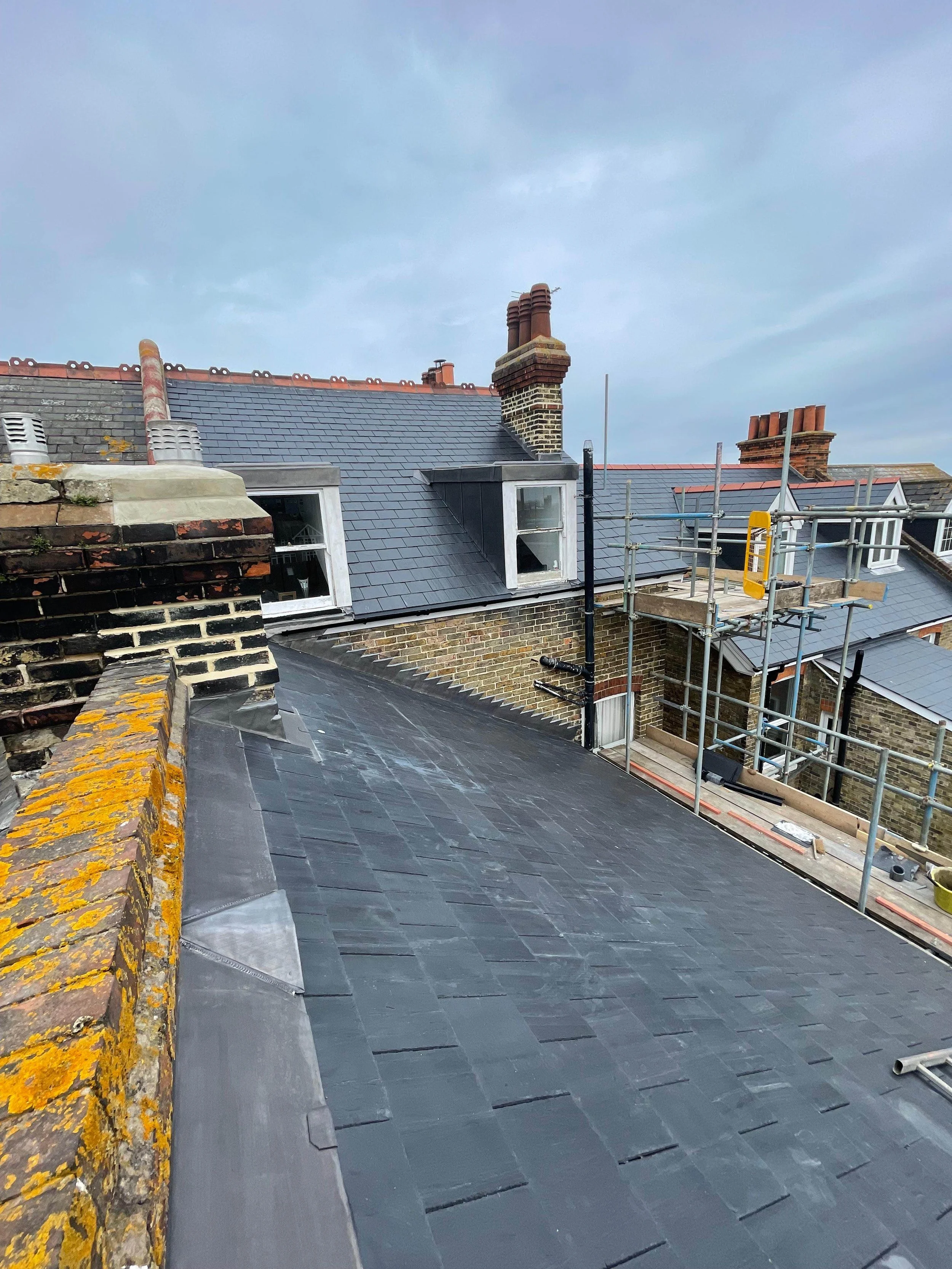 View of a rooftop under construction with dark slate tiles, scaffolding on the right, multiple chimneys, and cloudy sky.