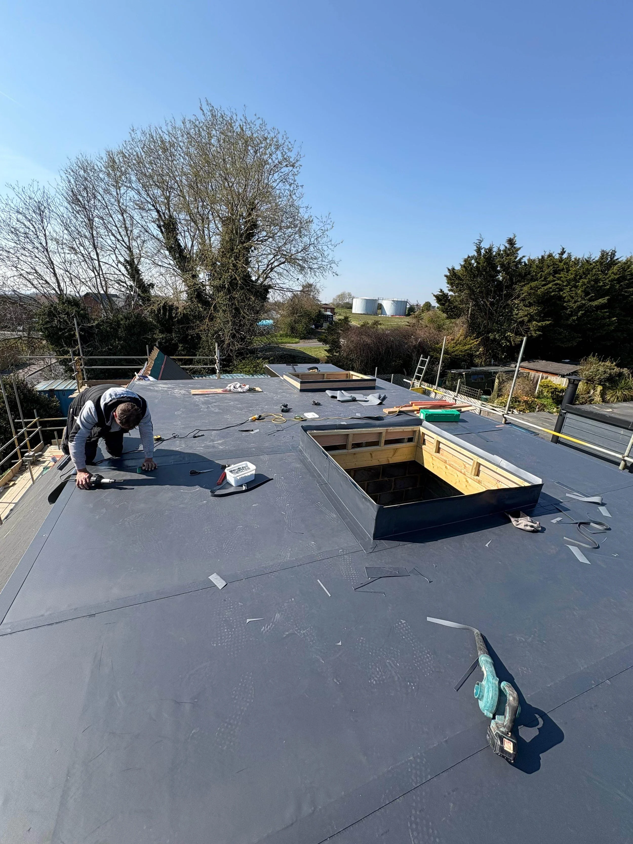 A worker installing a new roof on a building during the daytime, with construction tools and materials scattered on the roof surface.