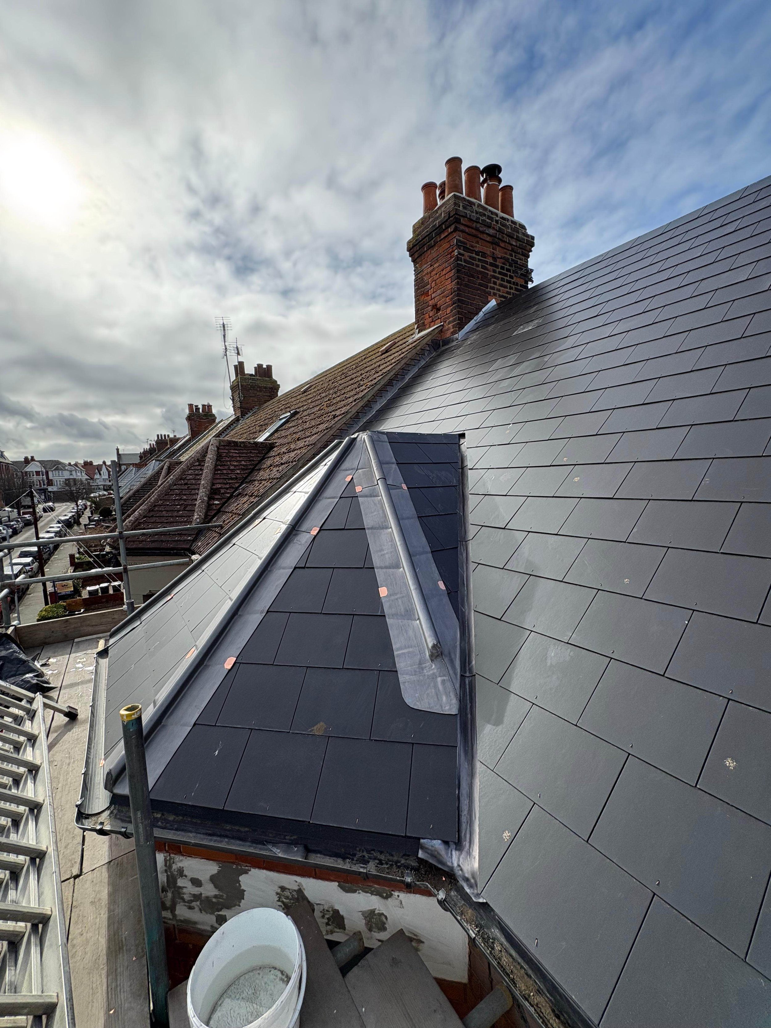 View of a roof undergoing installation or repair, with new black tiles and a metal flashing, and brick chimneys in the background under a partly cloudy sky.