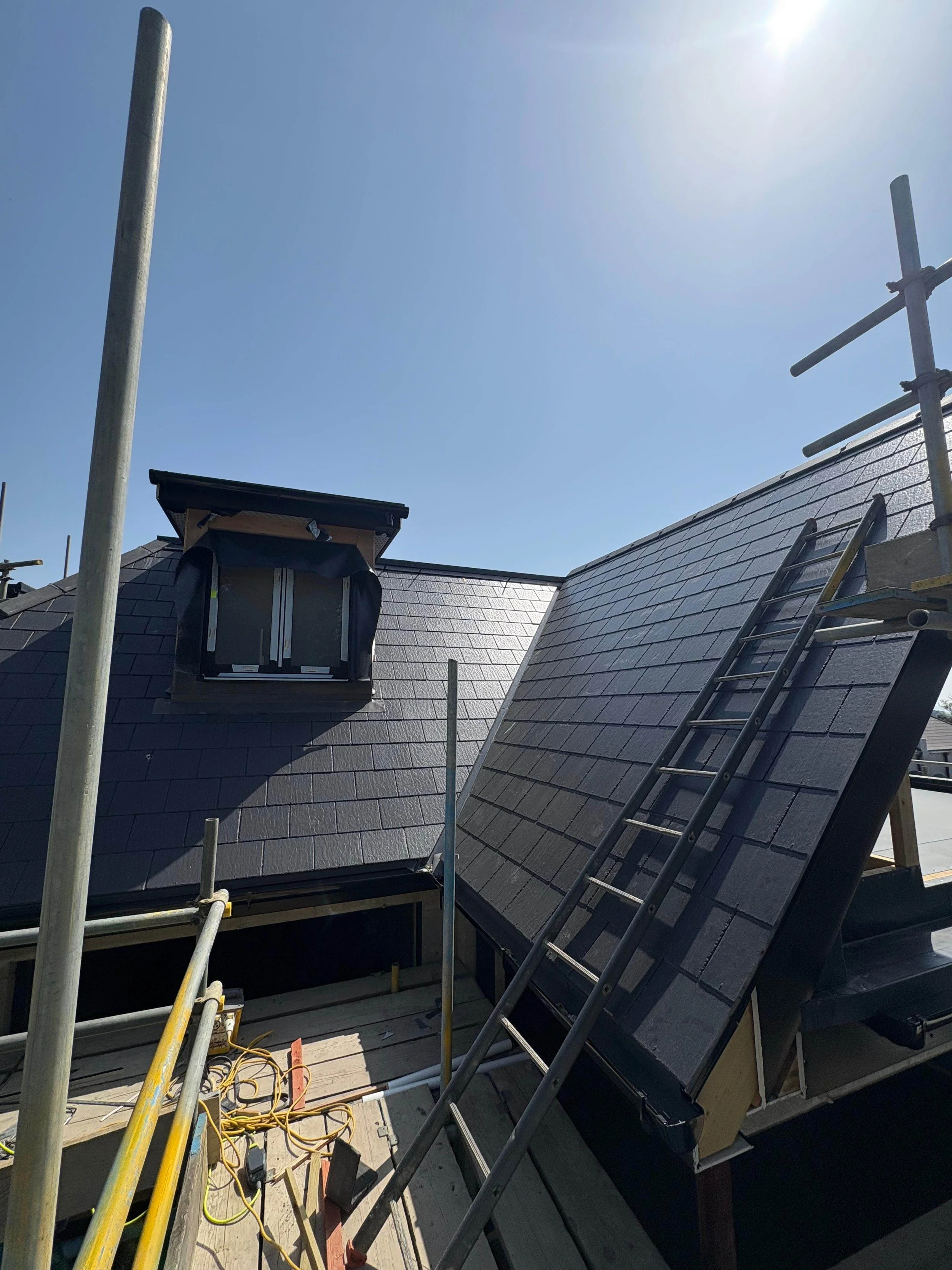 Roof renovation under construction with scaffolding, ladders, and solar shingles on a bright, sunny day.