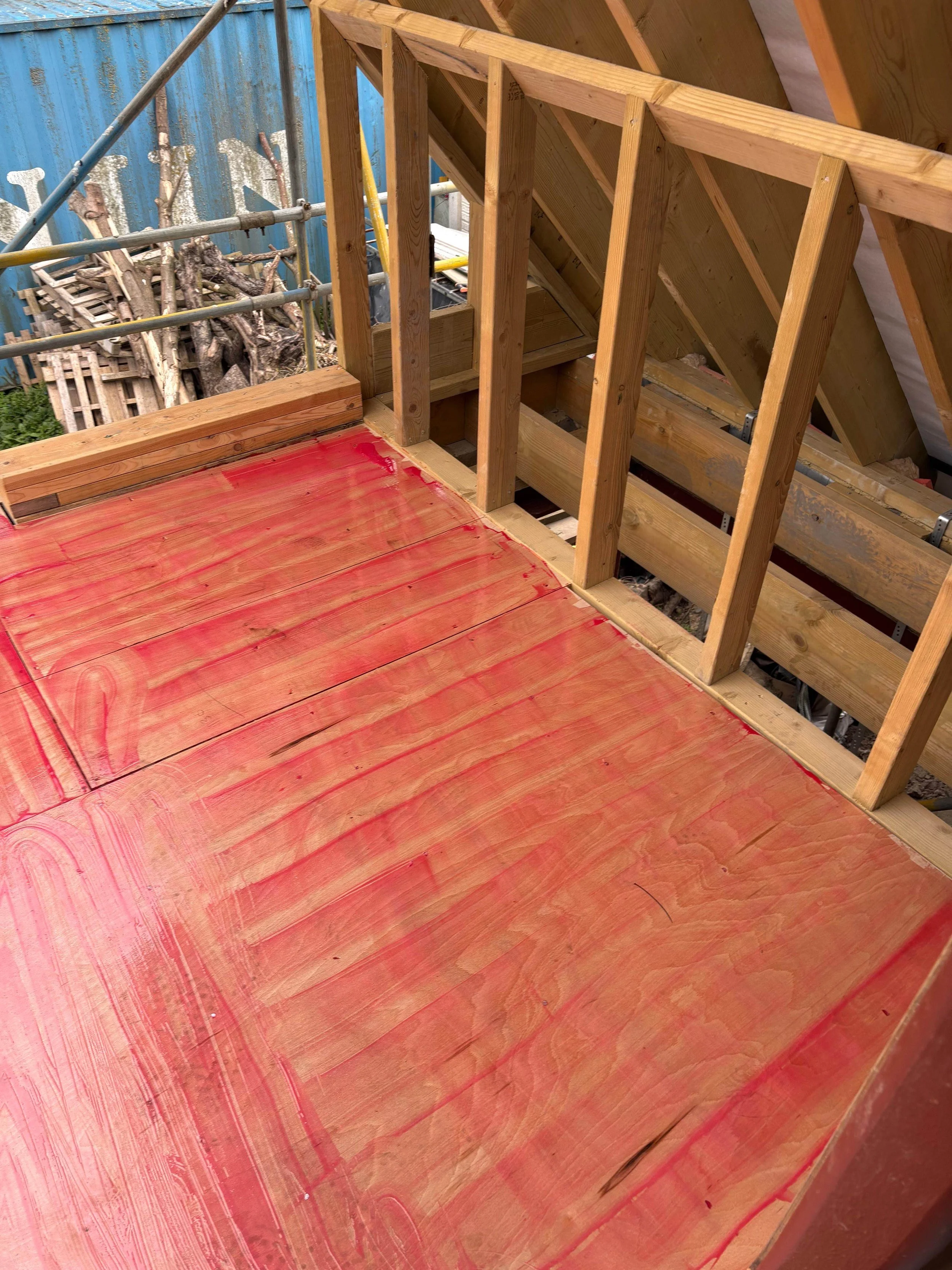 Wooden deck under construction with visible framing and partially installed red painted flooring. Construction tools and materials visible in the background.