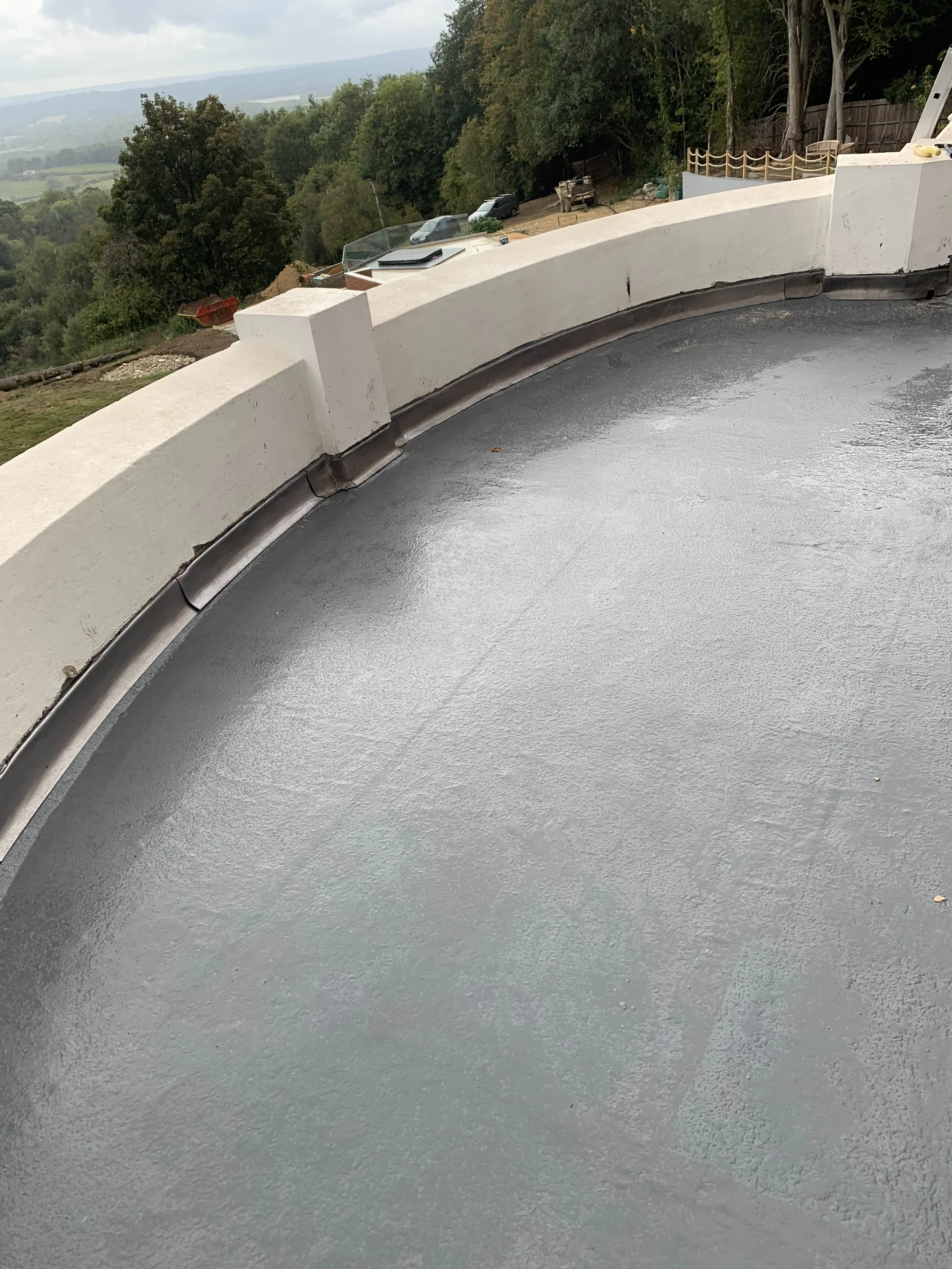 A curved concrete balcony floor with a white concrete railing, and a metal seam or joint along the edge. In the background, there are trees, parked cars, and a distant landscape with a cloudy sky.