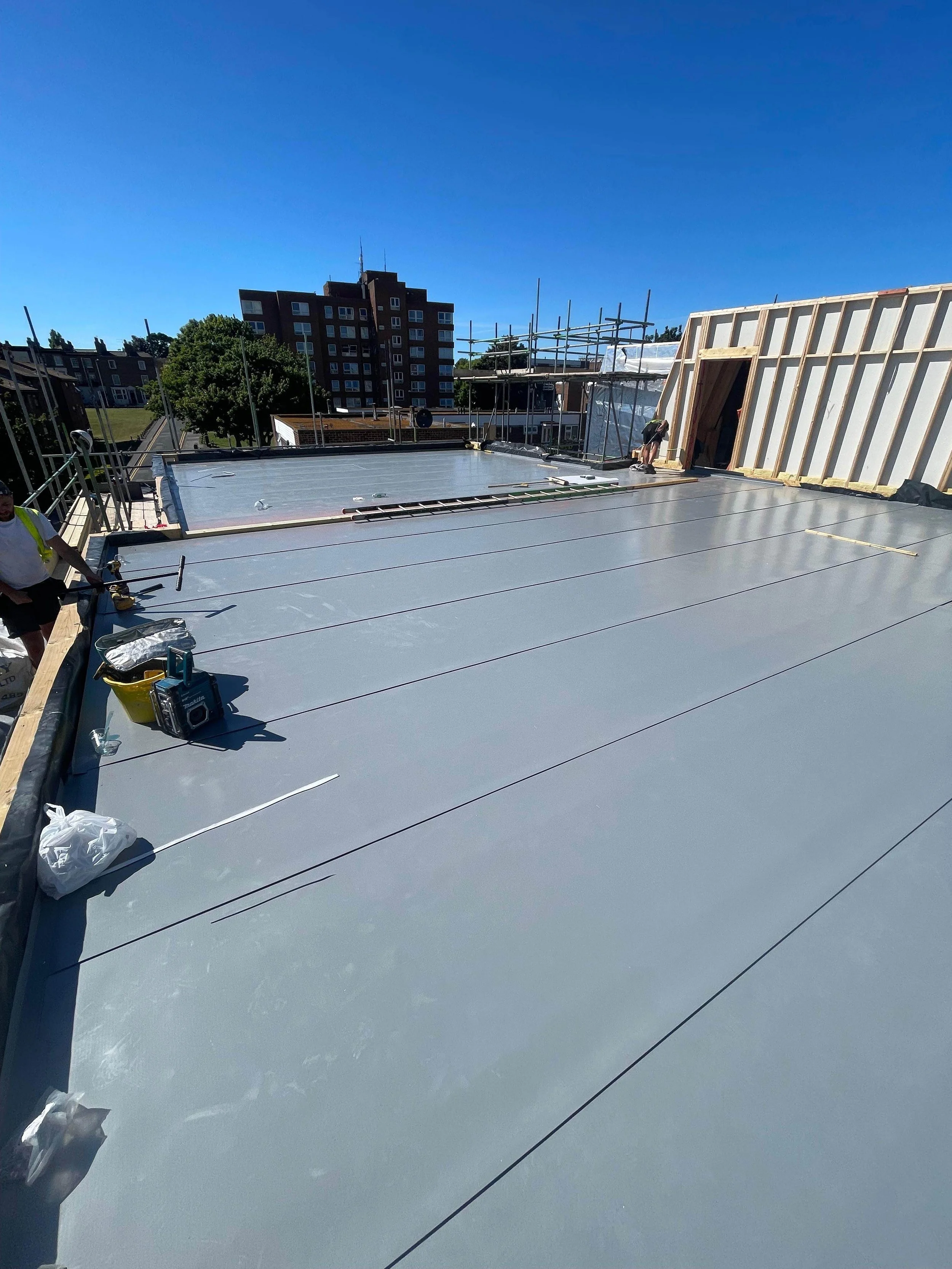 Construction workers installing a new roof with insulation and waterproofing on a building, with a cityscape of apartment buildings and trees in the background.