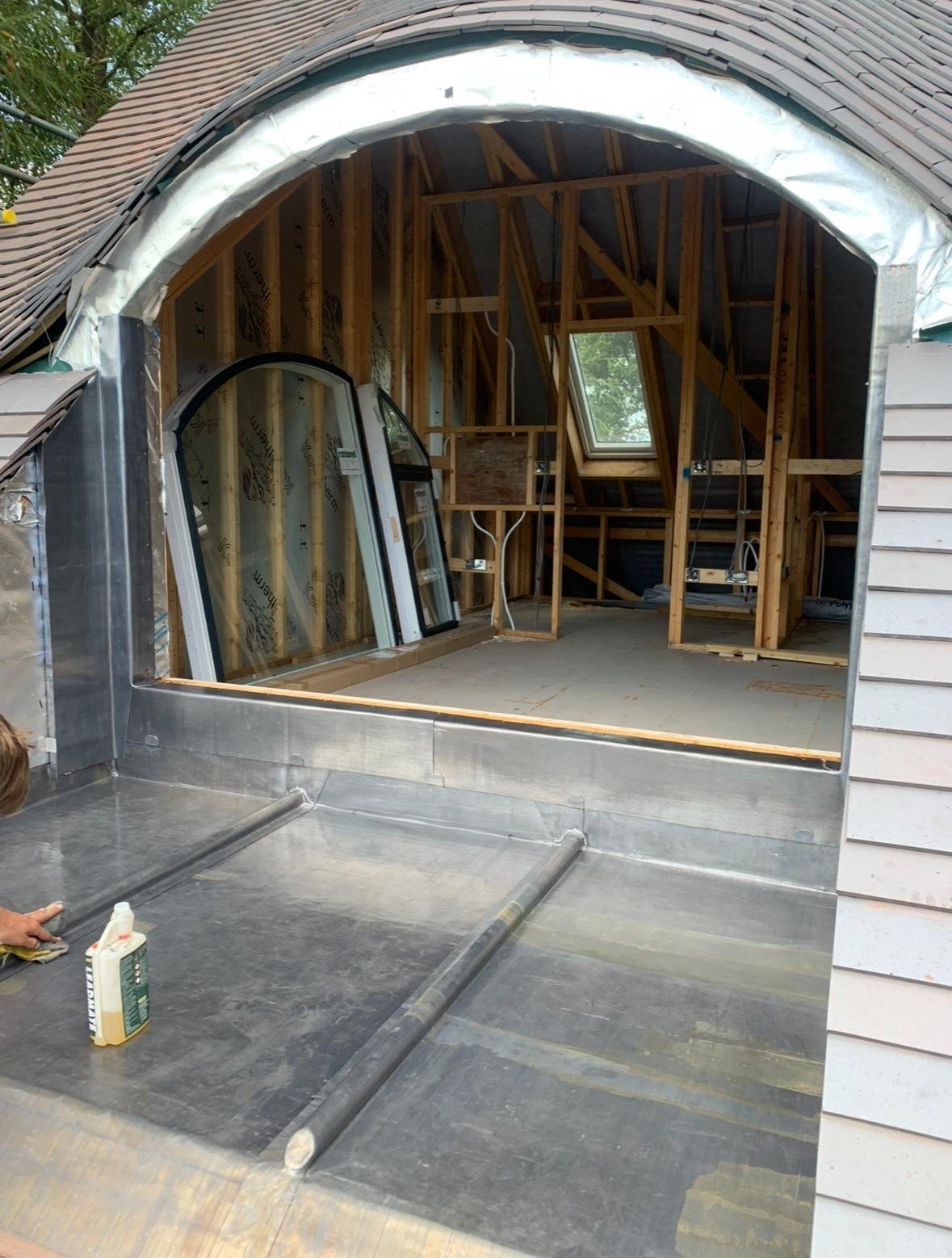 The image shows an unfinished interior space of a building under construction, with exposed wooden framing, a window, and construction materials. An opening in the building's wall reveals an exterior metallic surface and a partial view of a person wo