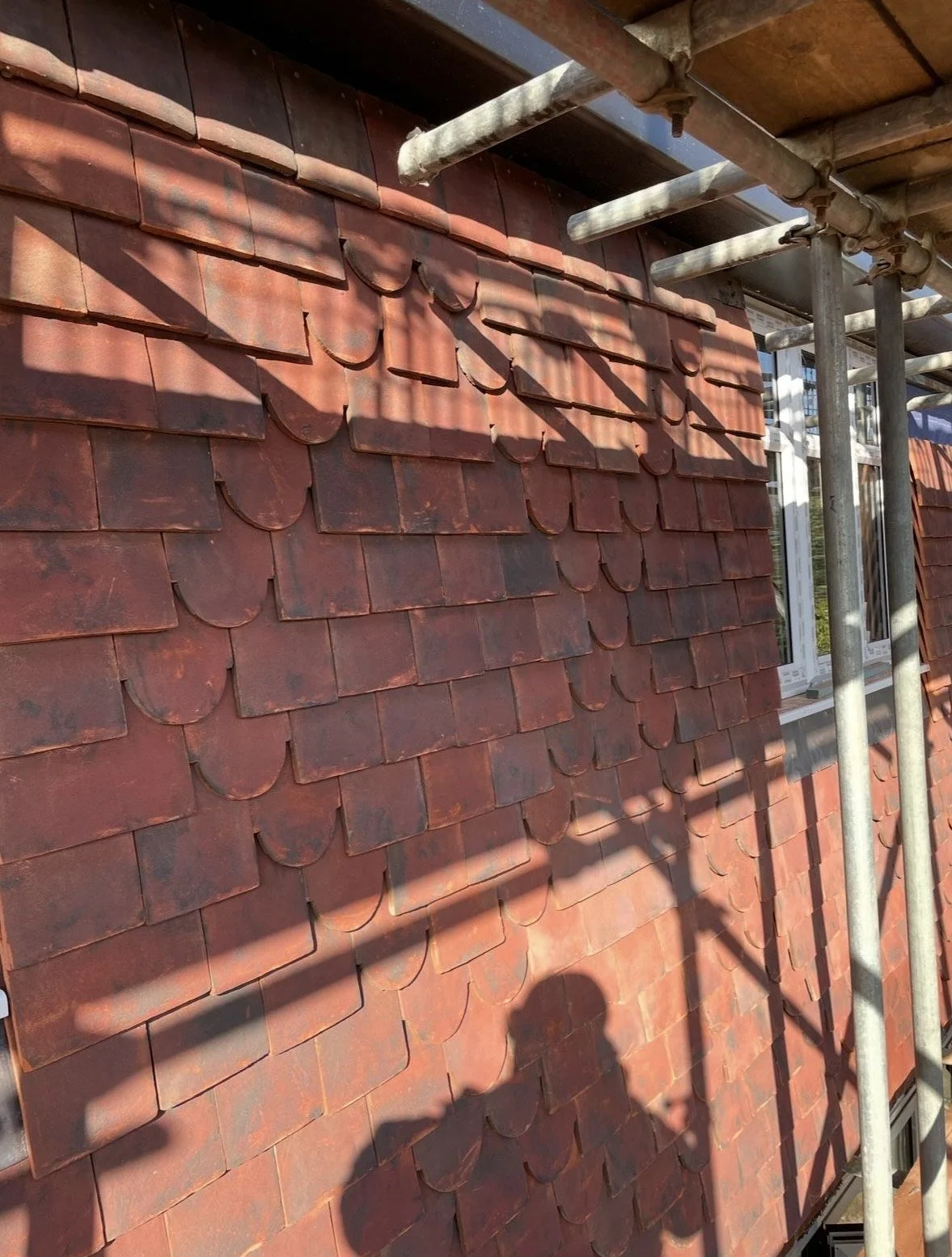 Red clay roof shingles on a building under construction or renovation, with scaffolding and shadows cast on the wall.
