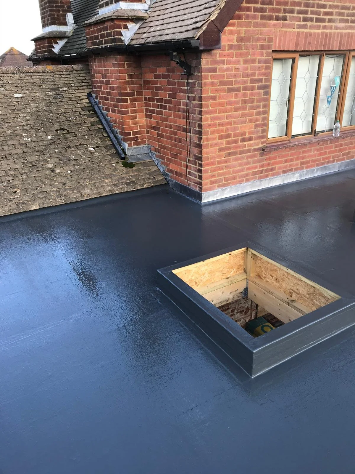 View of a flat roof with black waterproofing coating and a square opening with wooden framing, attached to a brick building with a sloped roof and a chimney.