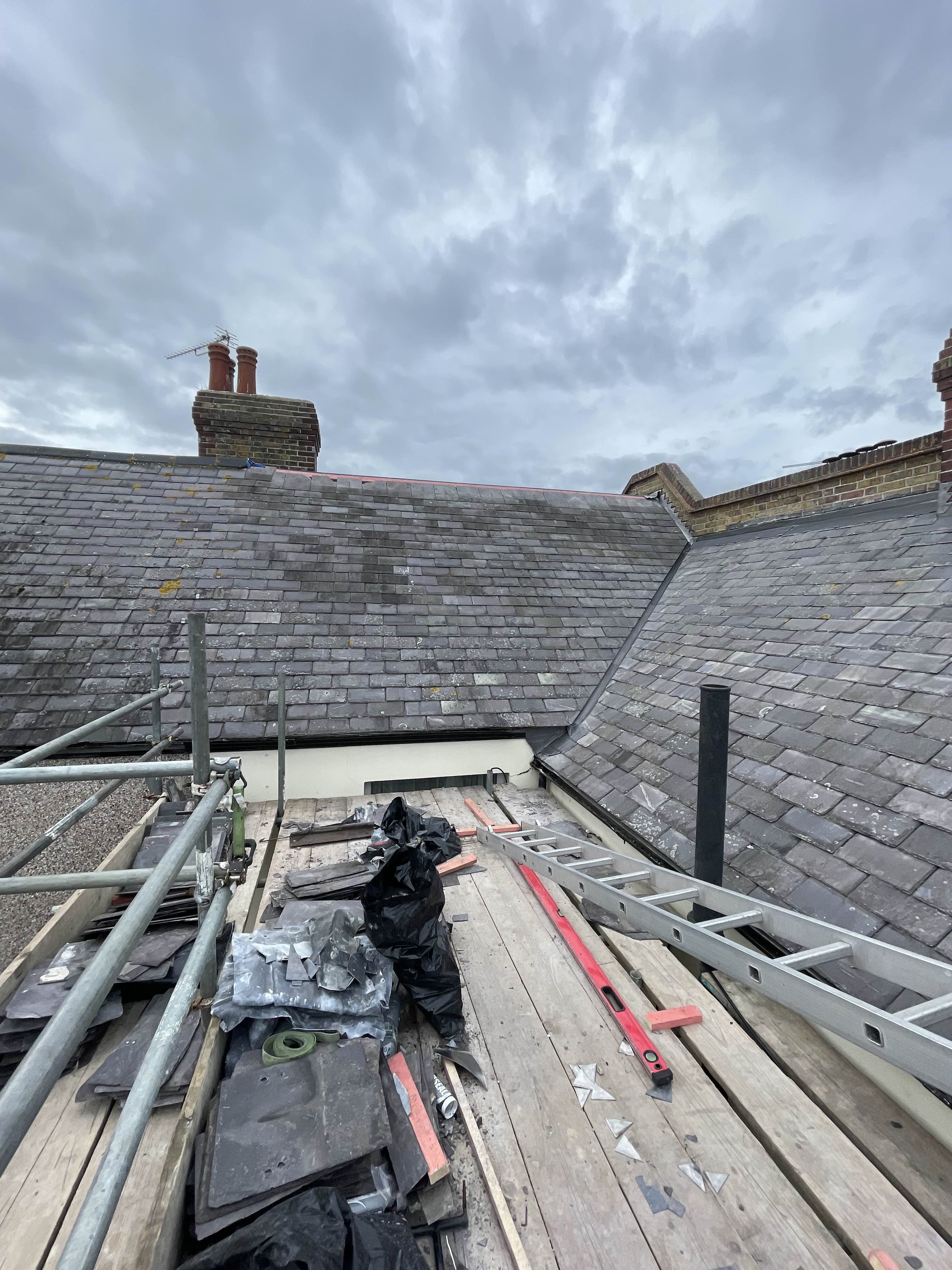 View of a rooftop under construction with various tools and materials, including a ladder, black bag, and tiles, with neighboring rooftops and a cloudy sky in the background.