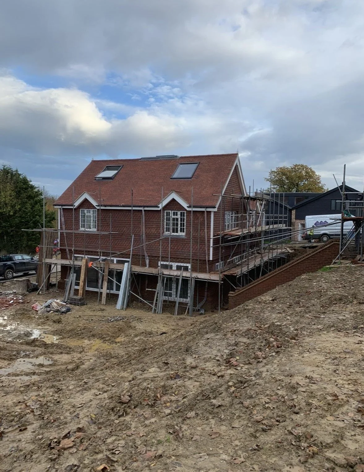 Construction site with a brick house surrounded by scaffolding and a dirt foreground, partly cloudy sky in the background.