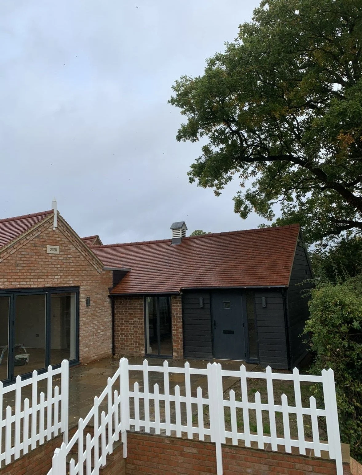 Exterior view of a house with brick and black siding, red tile roof, and white picket fence, with large tree and cloudy sky in background.