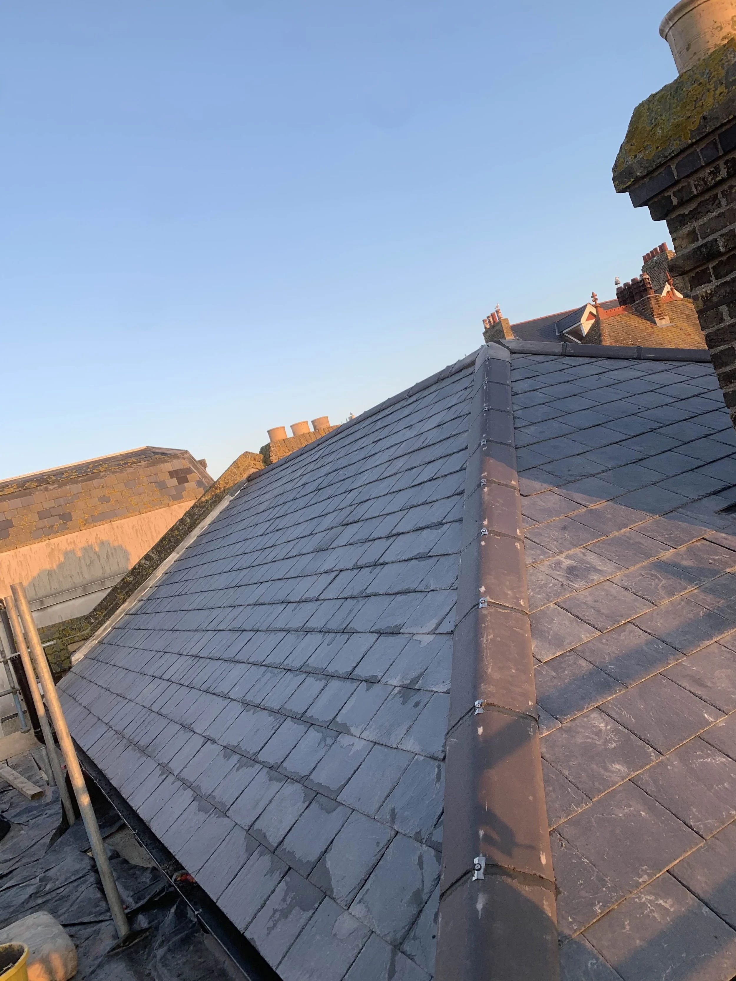 Close-up view of a residential roof with slate tiles and a ridge cap, under a clear blue sky.
