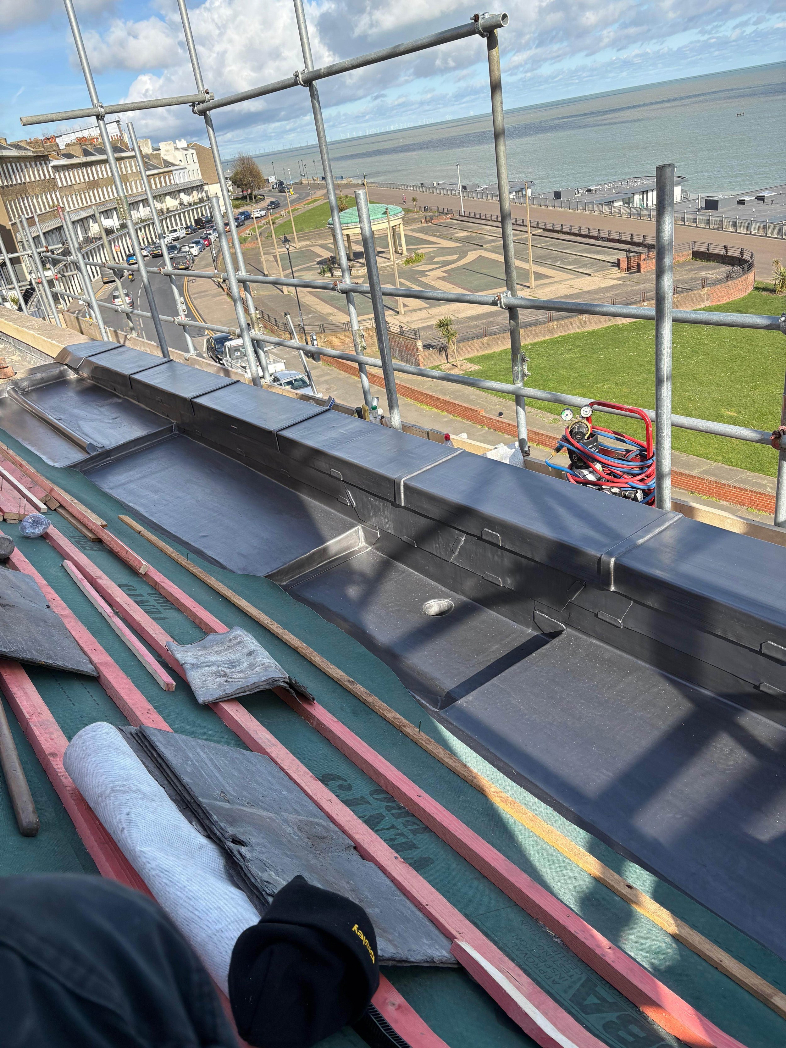 View from a rooftop under construction, showing roofing materials and scaffolding with a coastal park, beach, and ocean in the background.