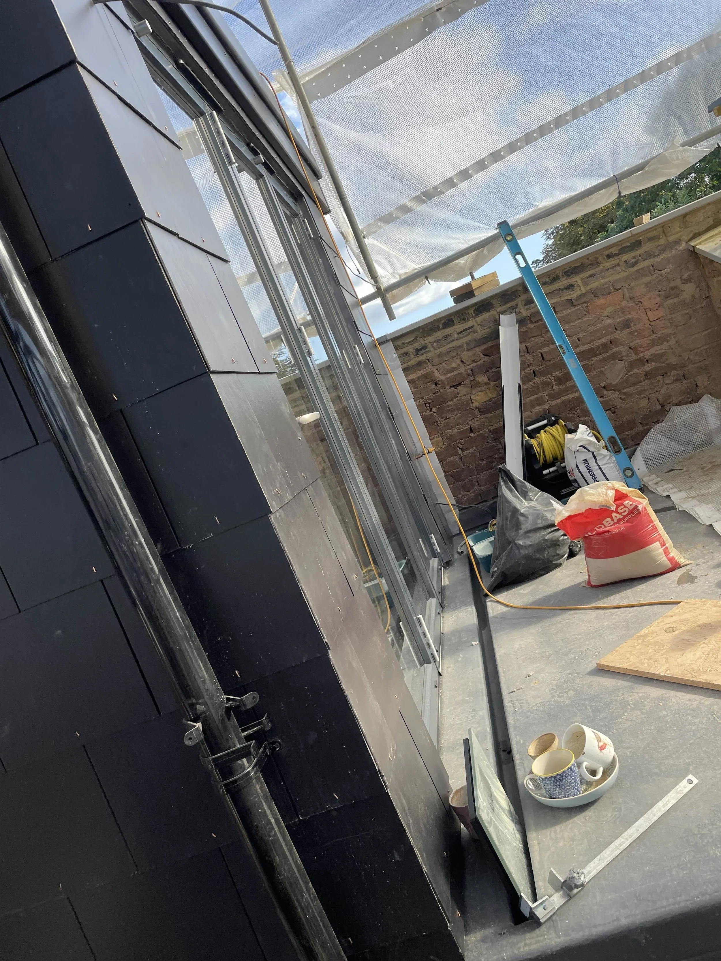 Construction site with black roof shingles, stacked window frames, a level, a bowl with cups, bags of construction materials, and a yellow and black hose reel on the floor, with a brick wall and partially covered roof in the background.