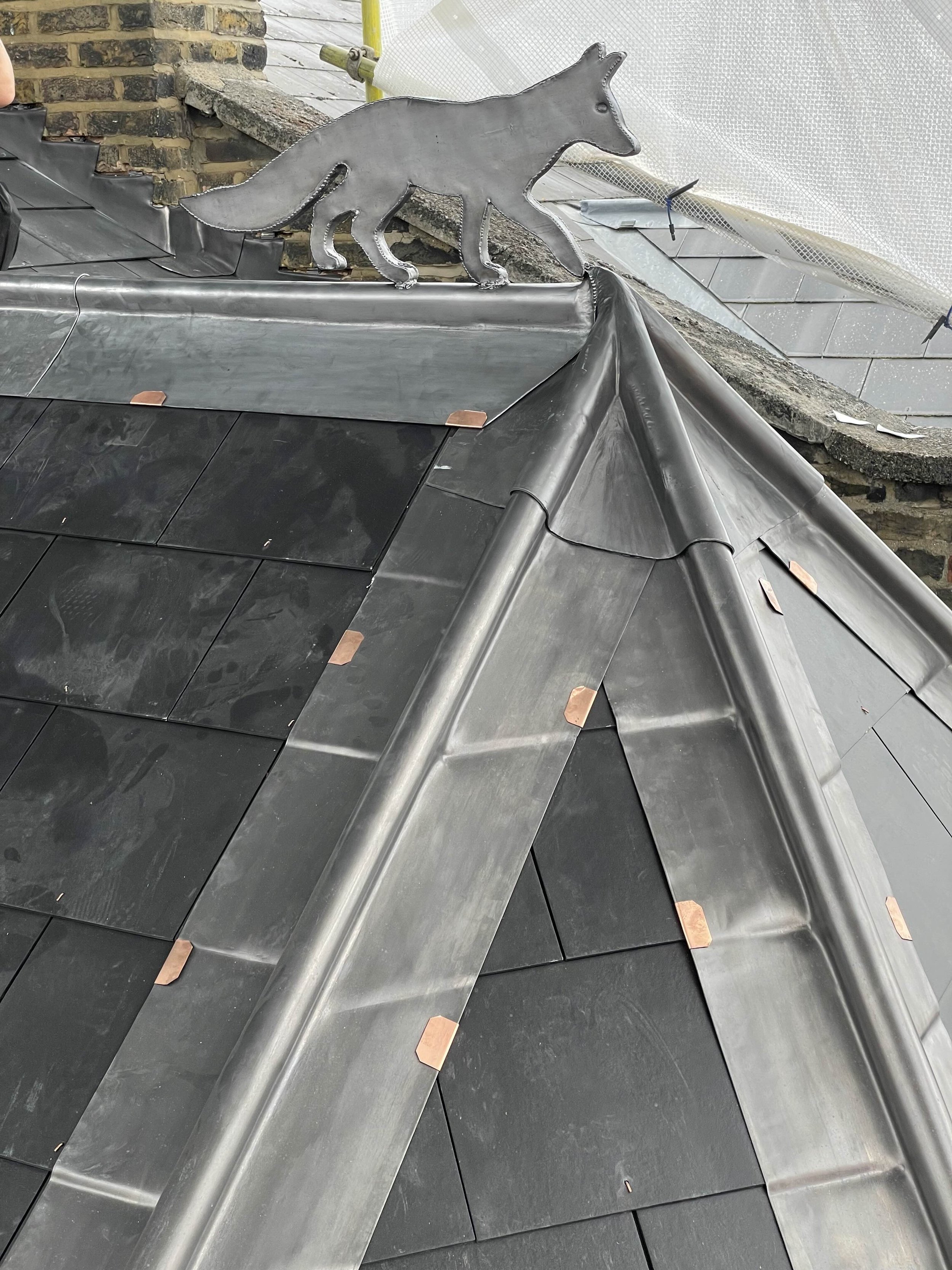Close-up of a metal roof with black tiles, copper fasteners, and a decorative metal fox on the ridge.