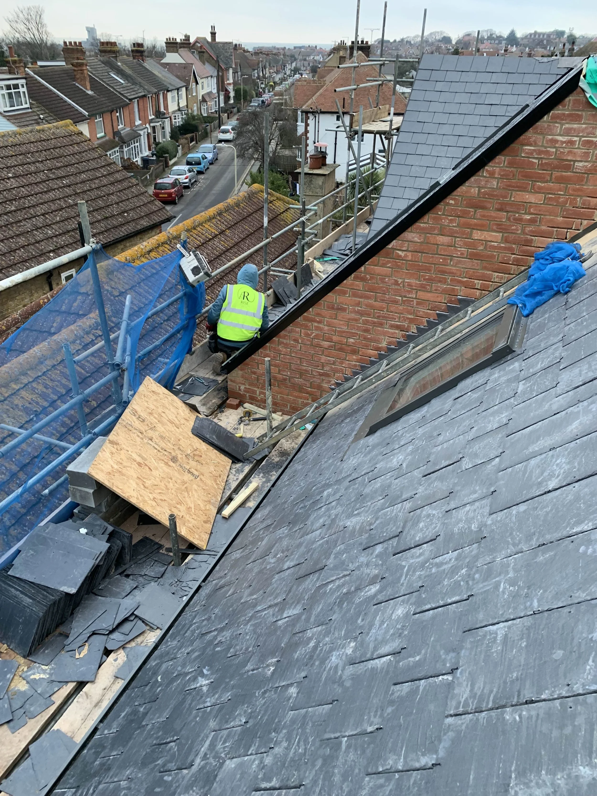 A worker in a yellow safety vest and blue hoodie is installing or repairing roofing on a house. The roof has dark slate tiles, and there is scaffolding with a blue safety net around the perimeter. Construction materials and tools are nearby on the ro