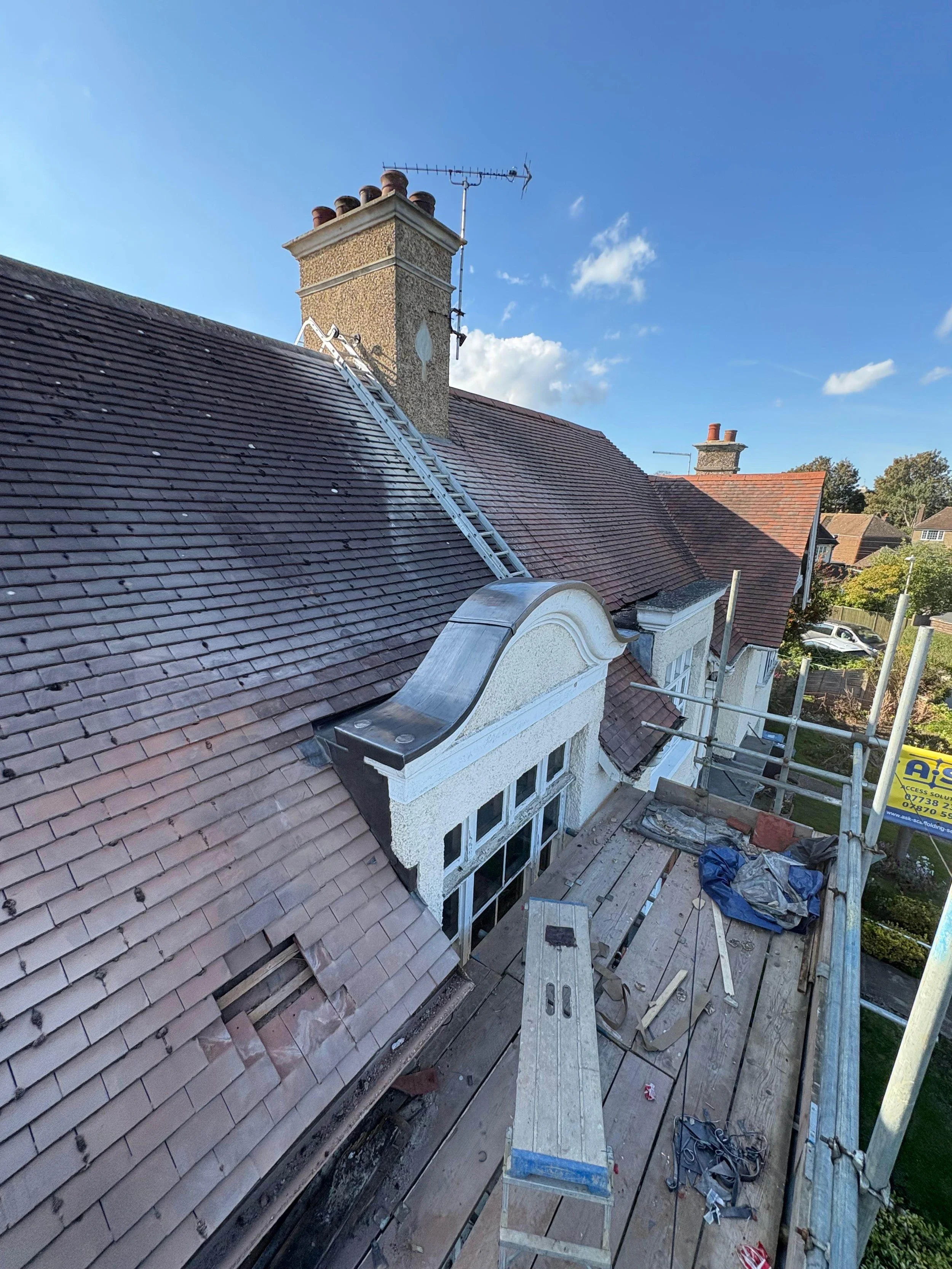 View from a scaffolded rooftop showing a chimney, roof tiles, scaffolding, and building materials under a partly cloudy sky.
