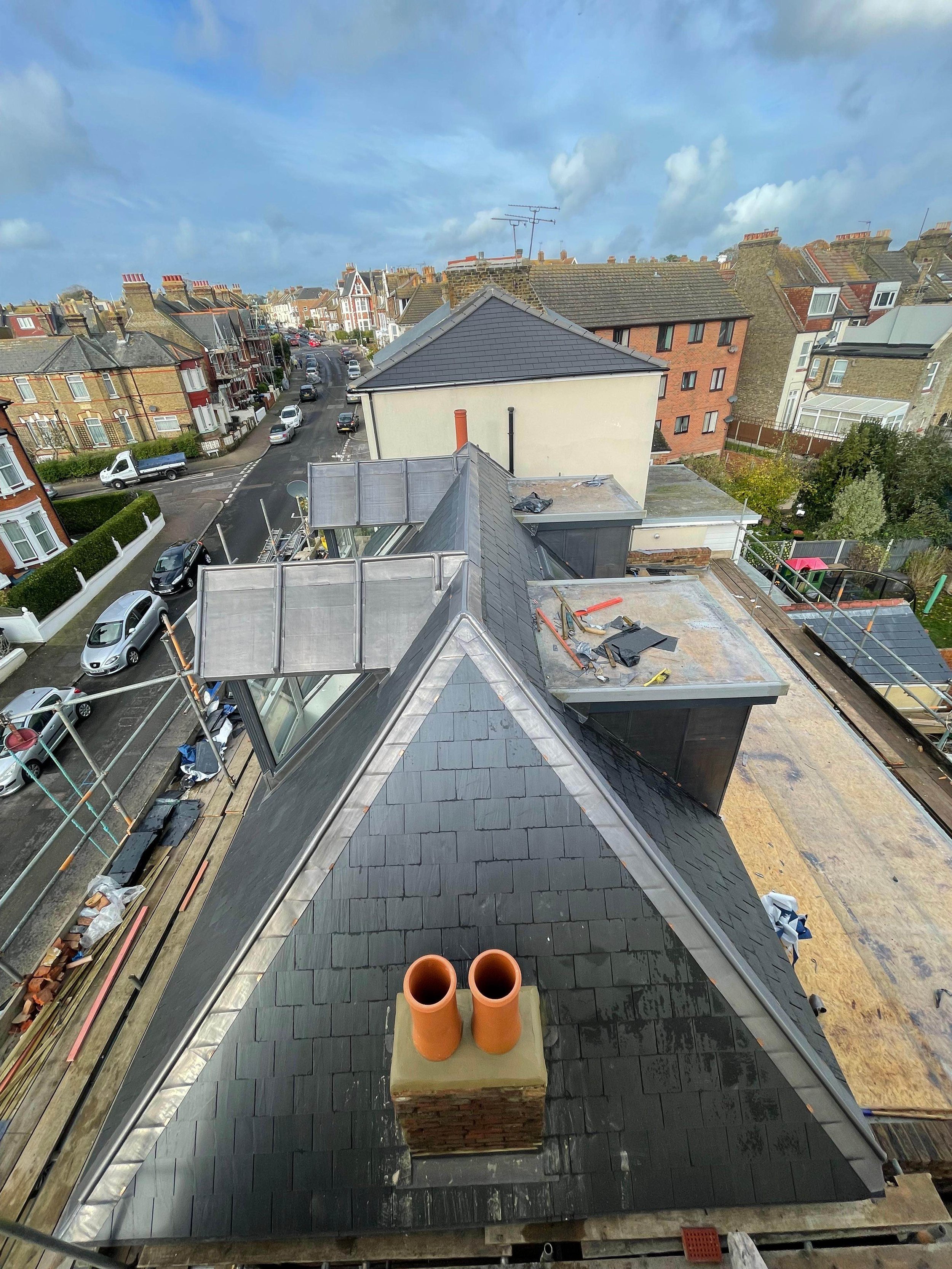 A rooftop under construction with black shingles, orange chimney pots, and scaffolding, overlooking a residential street with cars and houses.