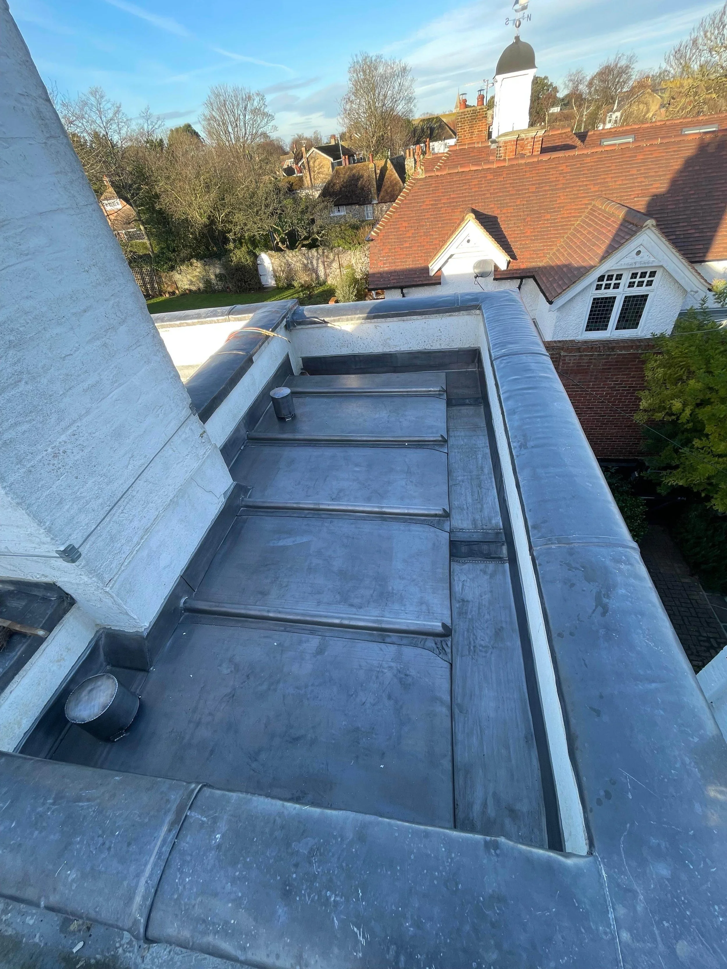 View from a rooftop showing a flat roof with a chimney, drain pipes, and a background of neighboring houses with red roofs, trees, and clear sky.