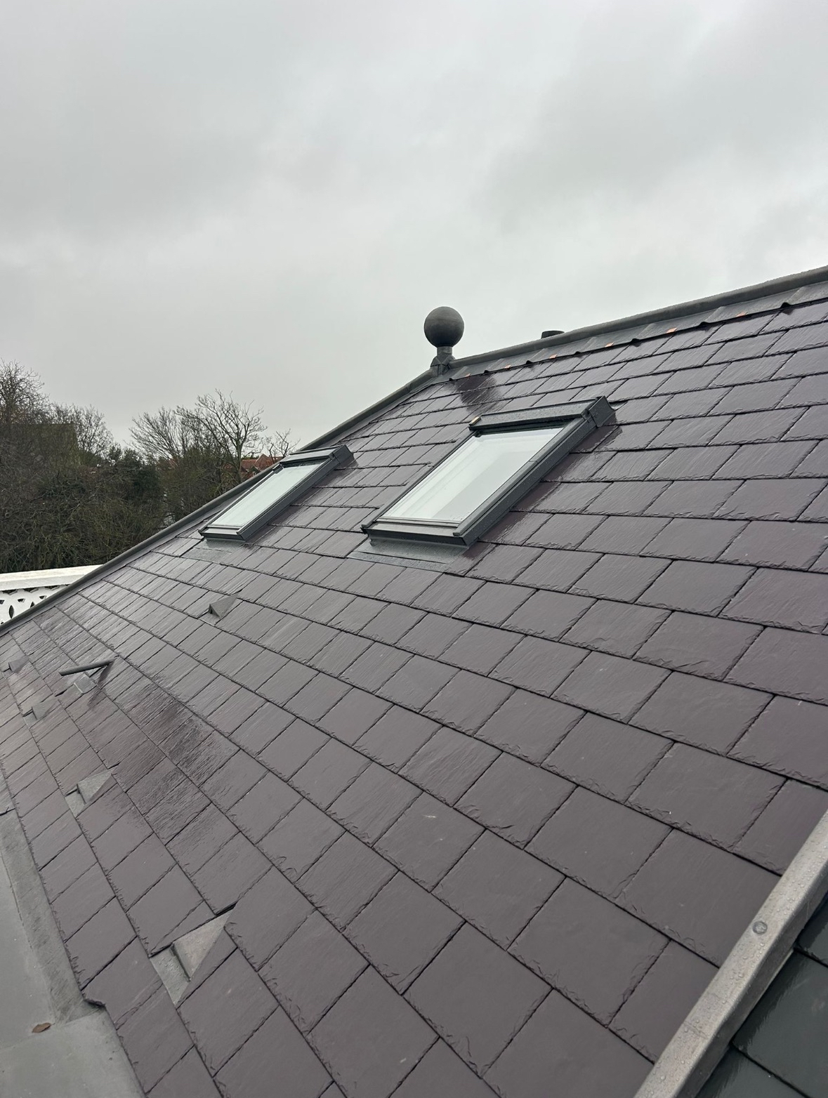 The image shows a gray tiled roof with two skylights and a black spherical finial on top, under an overcast sky.