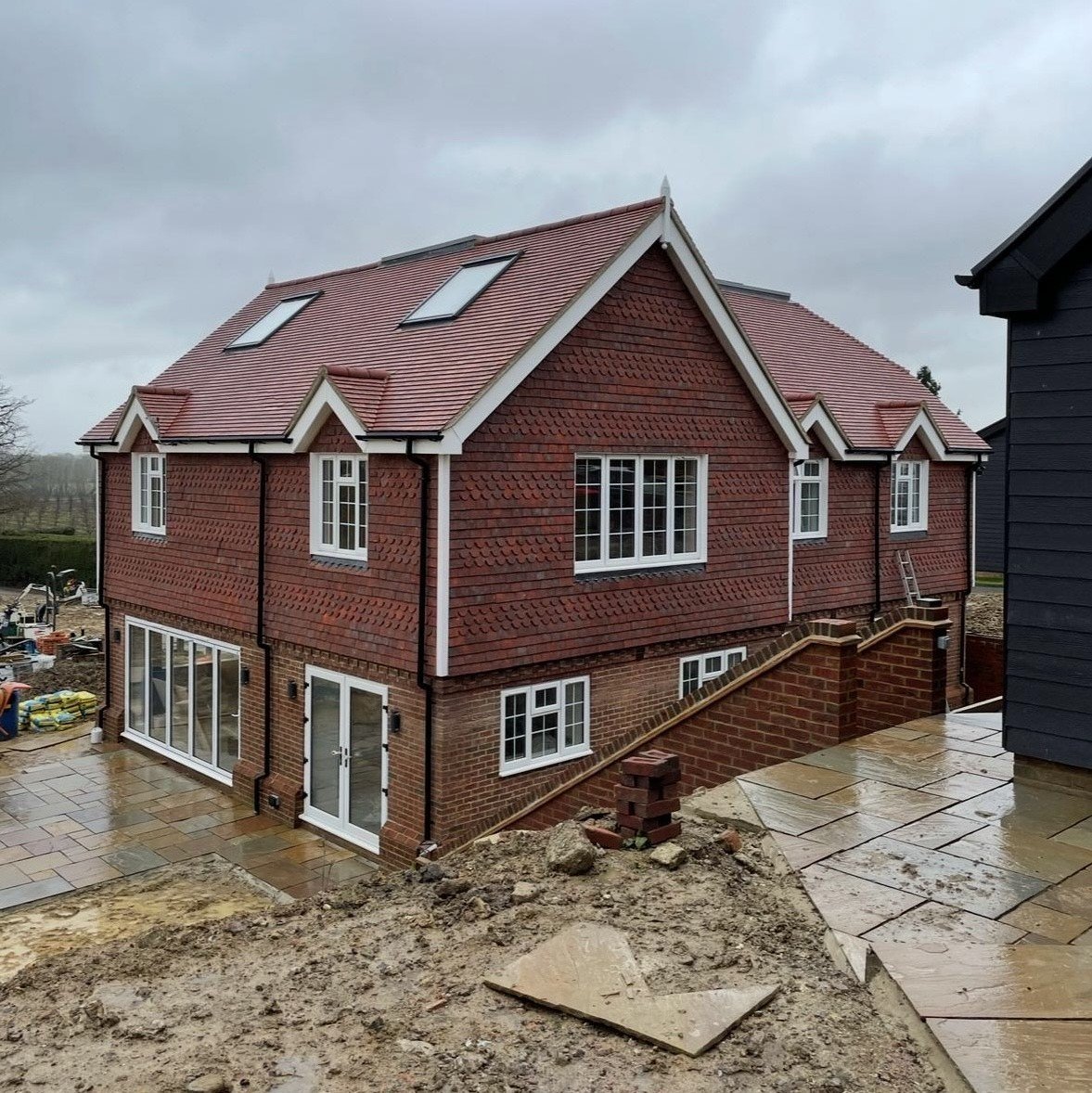 Newly built brick and shingle house with large windows, a sloped roof with skylights, and a stone-paved patio on a cloudy day.