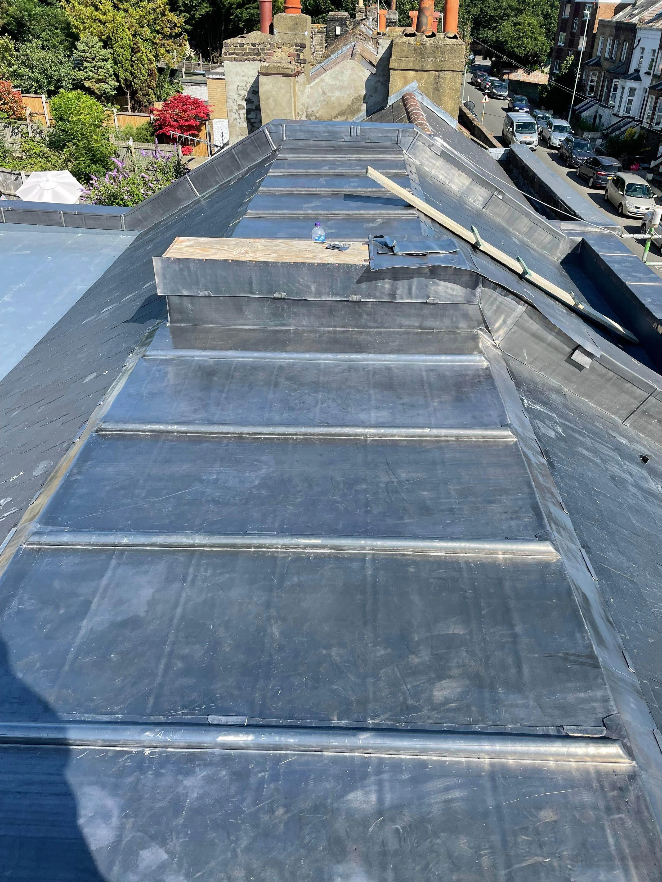 View of a metal roof under construction with some wooden planks and tools on it, residential area with houses, parked cars, and trees in the background.