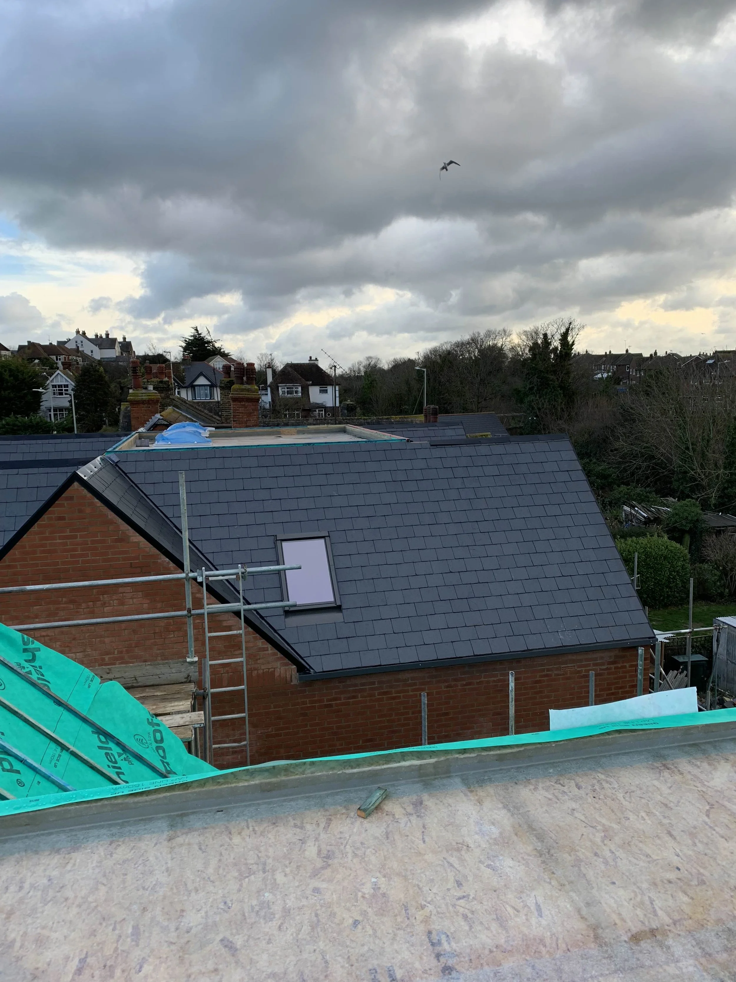 View of rooftops under a cloudy sky, with a bird flying above. The foreground shows a construction or renovation site with scaffolding and building materials.