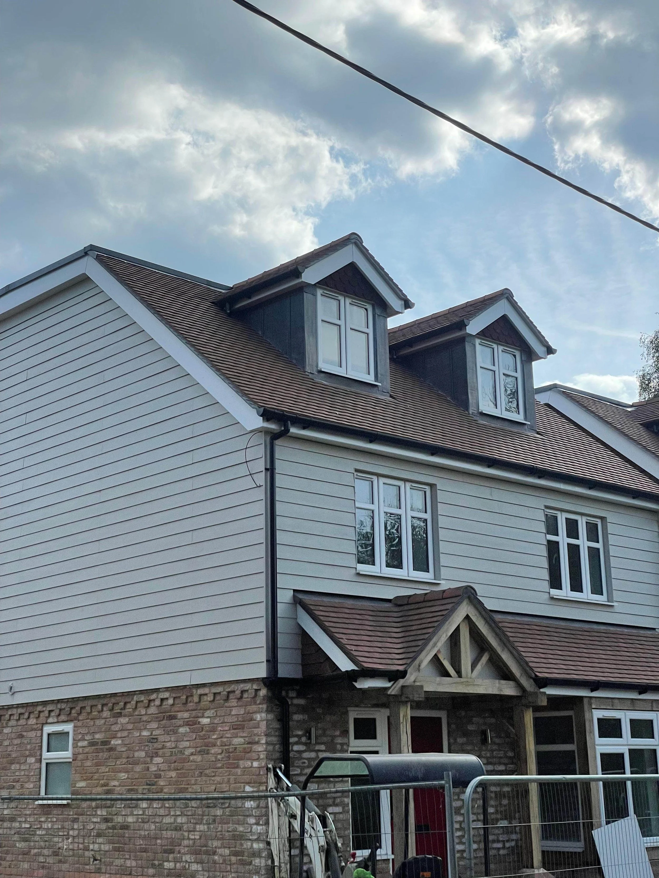A two-story house with cream-colored vinyl siding, a brick lower level, and three dormer windows on the roof. The sky is partly cloudy, and there's a power line above the house.