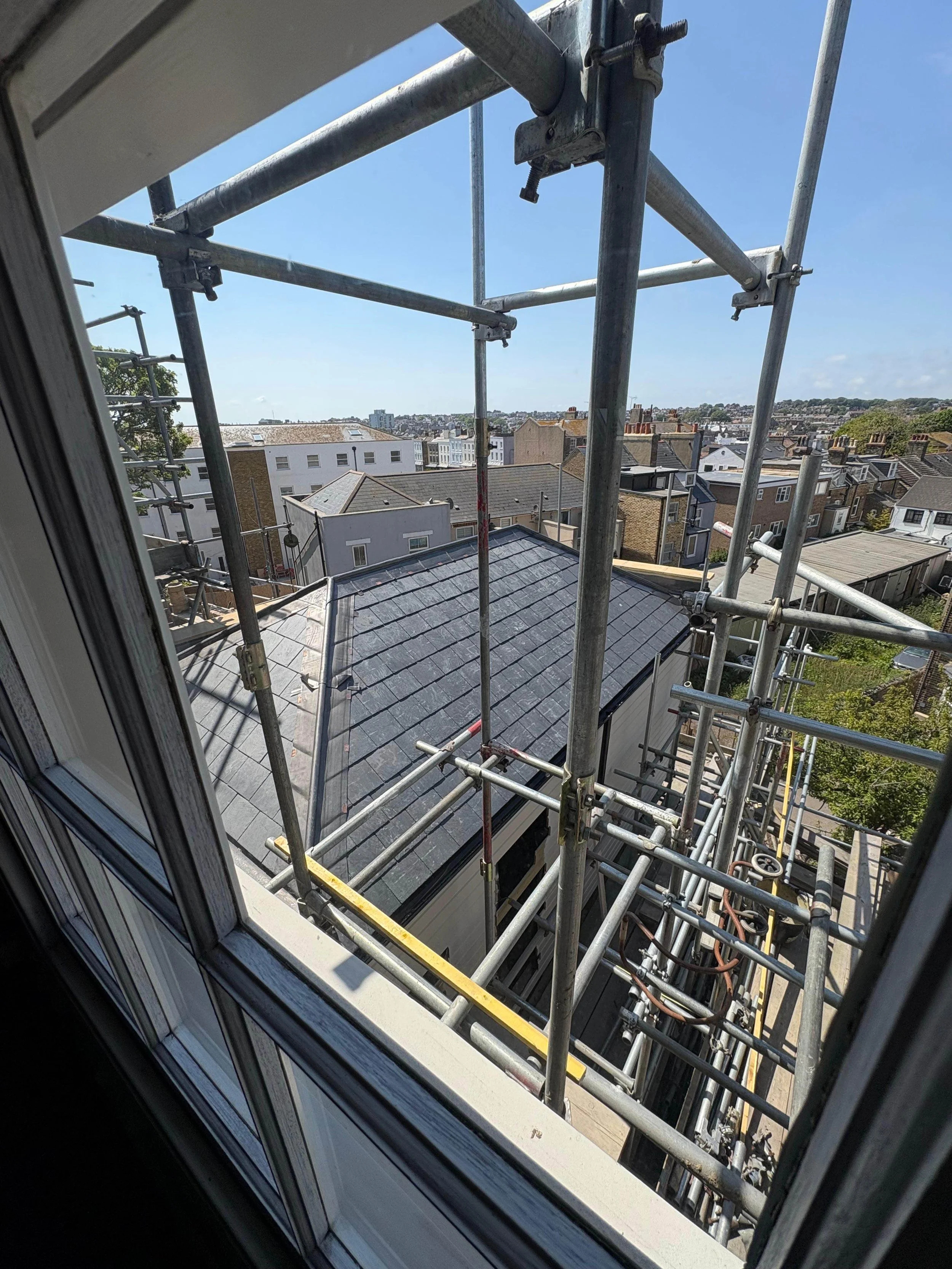 View from a window showing a rooftop and metal scaffolding around a building on a sunny day with a clear blue sky. The cityscape with multiple houses and buildings is visible in the background.