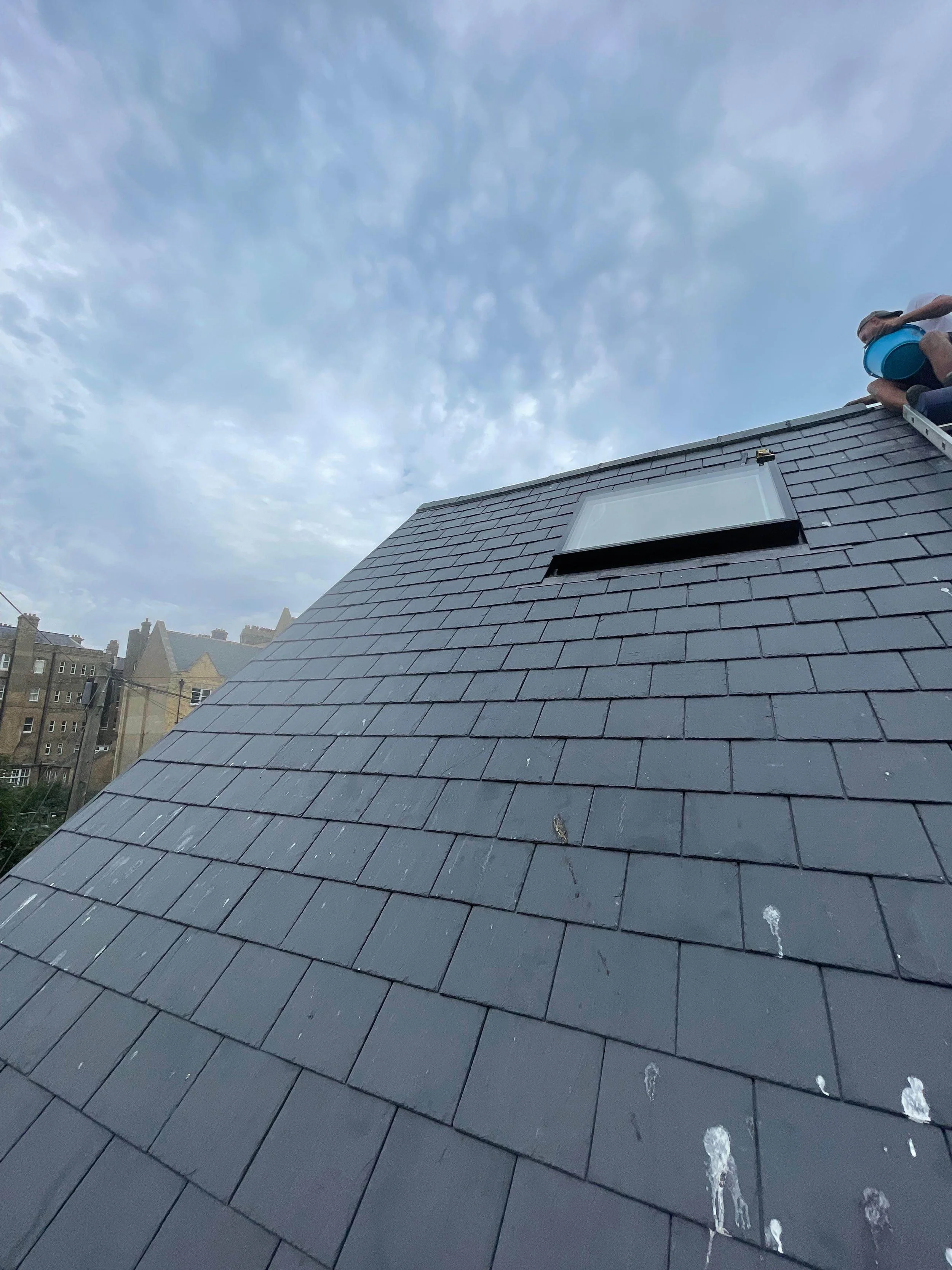 Close-up of a steep rooftop with gray shingles, a small window, and a worker sitting on the edge holding a blue container, against a partly cloudy sky.