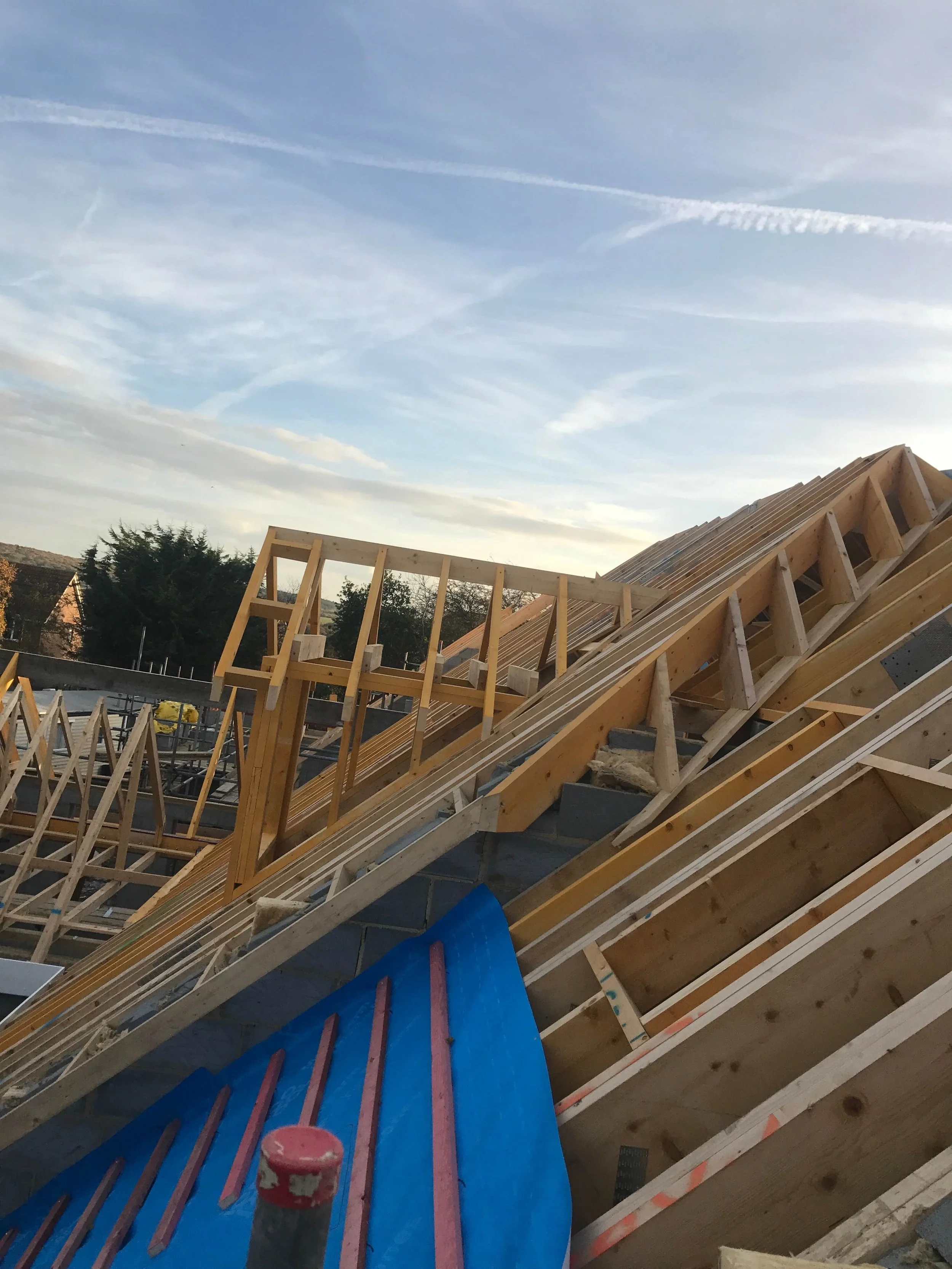 Construction site with wooden framework for a roof, blue tarp, and partially assembled roof trusses under a blue sky with wispy clouds.