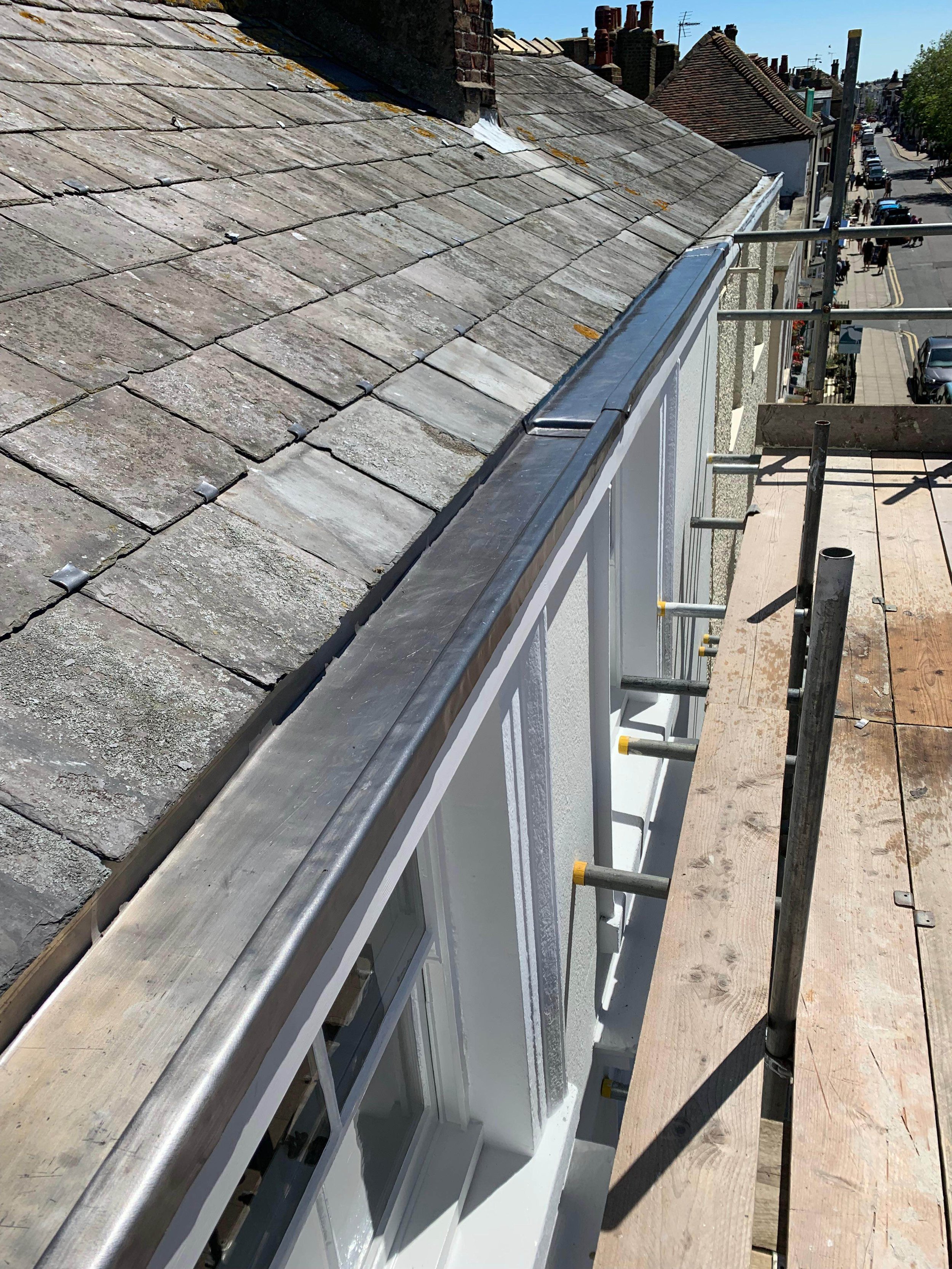 View from a rooftop construction site showing a wood scaffold platform, metal railing, and a slate-tiled roof overlooking a street with cars and pedestrians.