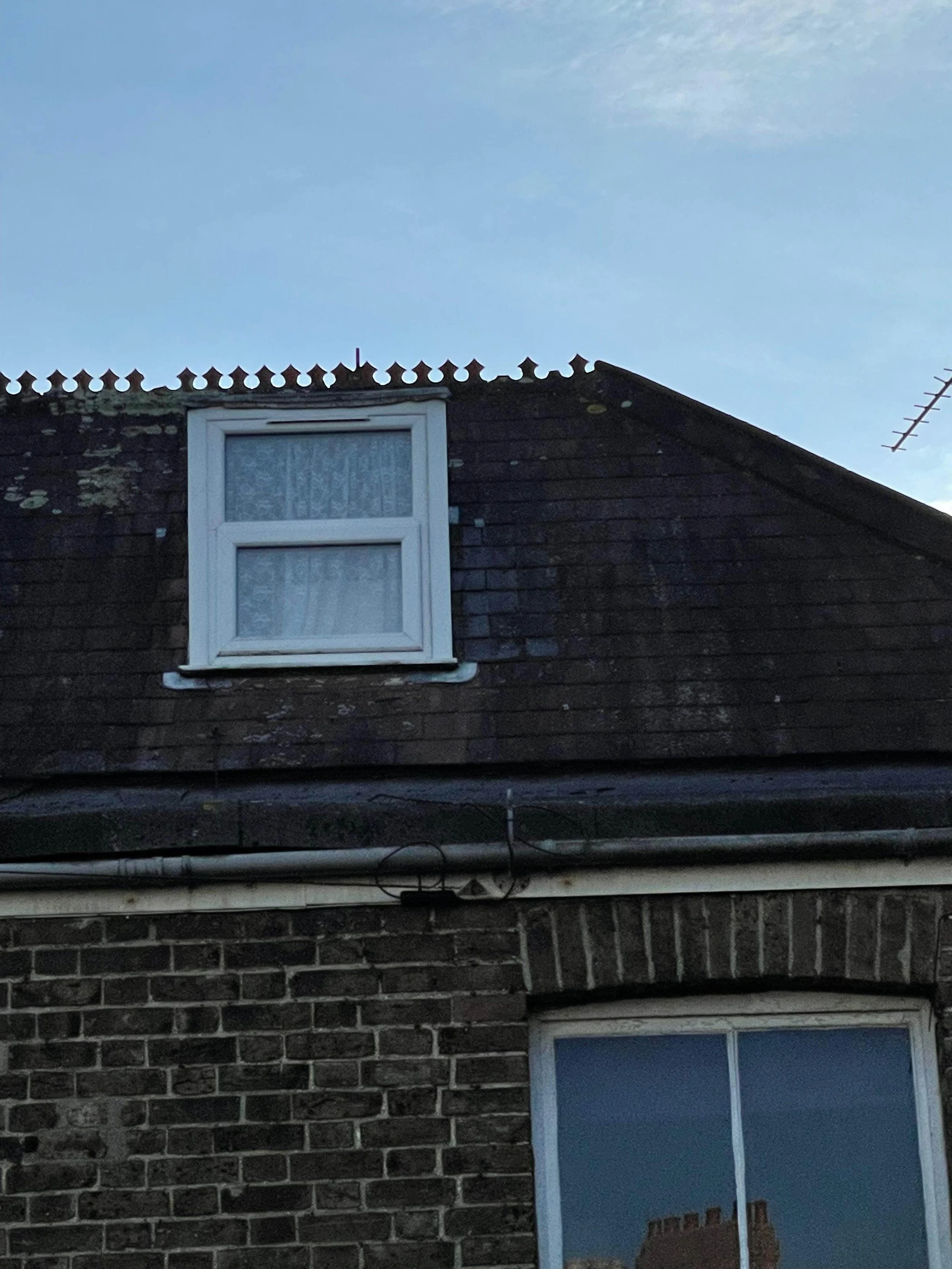 The upper part of a brick house with a single window on the second floor, weathered wooden siding, and a clear blue sky. The window has a white frame and lace curtains, with some moss or algae on the siding. A gutter runs across the middle of the ima