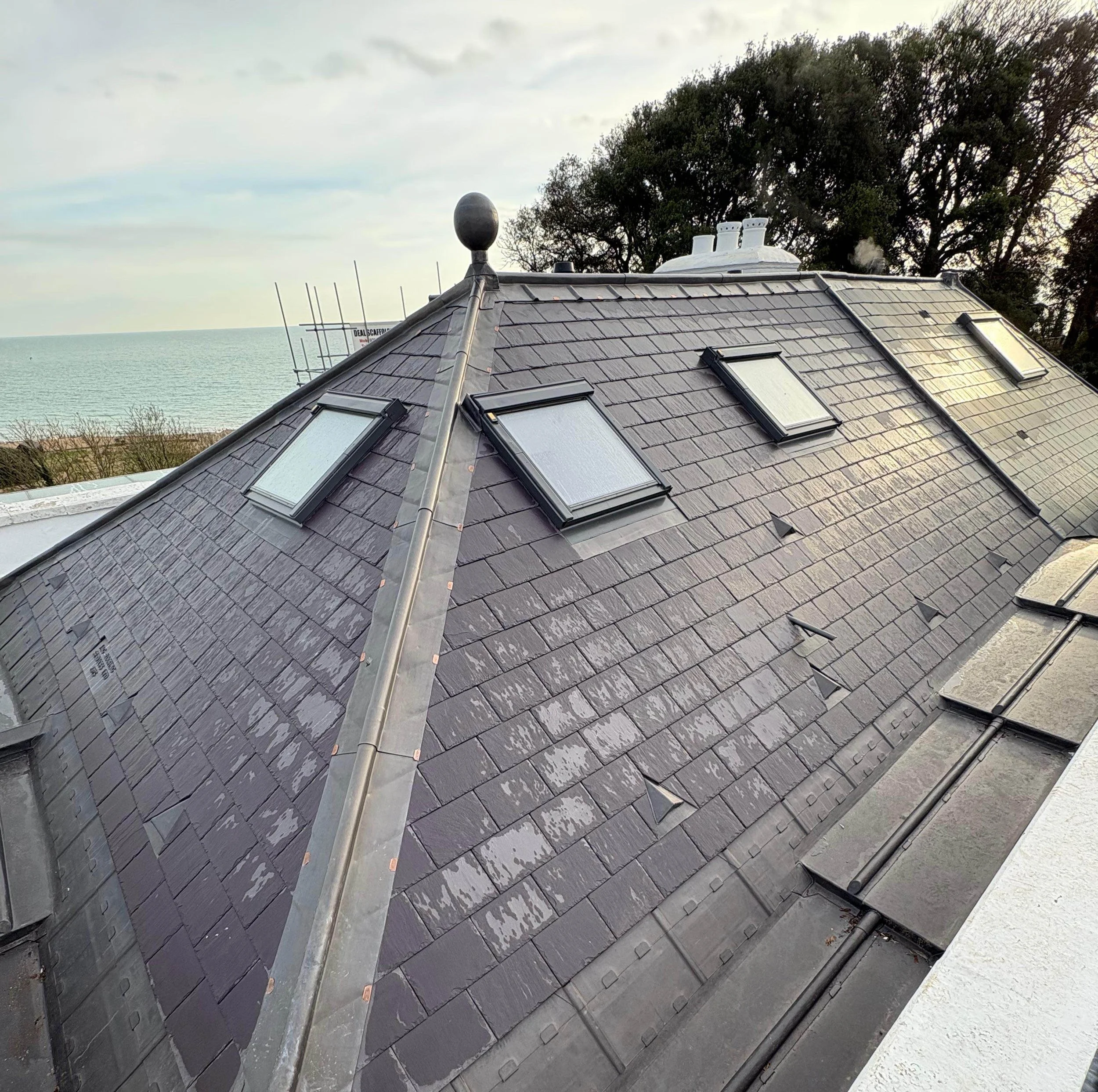 Residential roof with multiple skylights, a metal ridge vent, and a chimney near the top, overlooking trees and a body of water in the distance.
