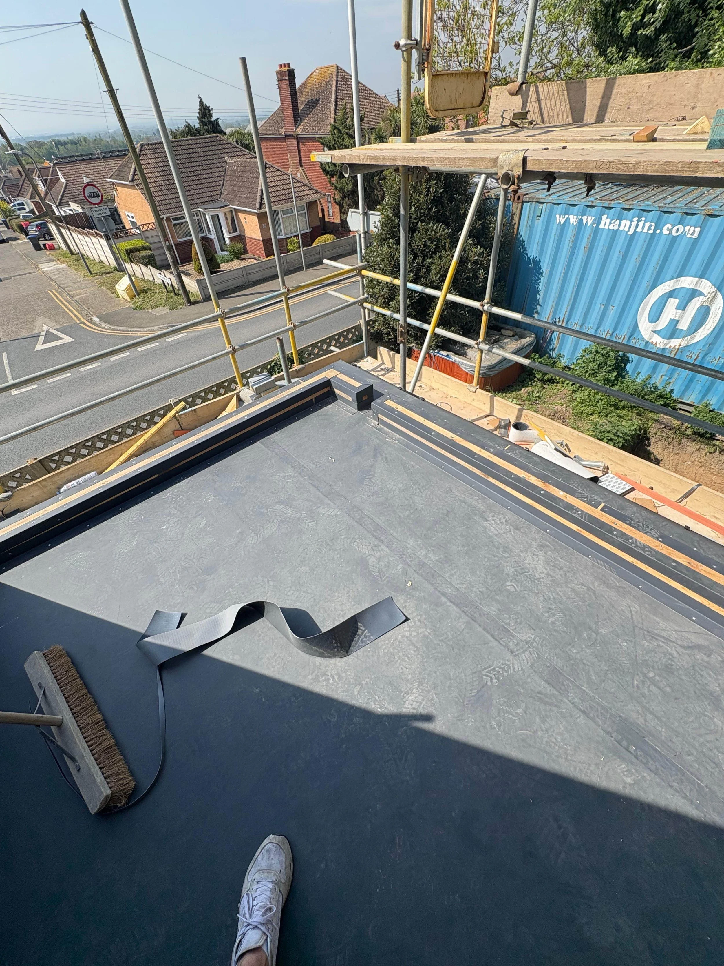 View from a high construction site showing a flat roof with construction materials, a broom, and part of a person's foot in a white sneaker. The background features a residential neighborhood with houses, trees, and a construction site with scaffoldi