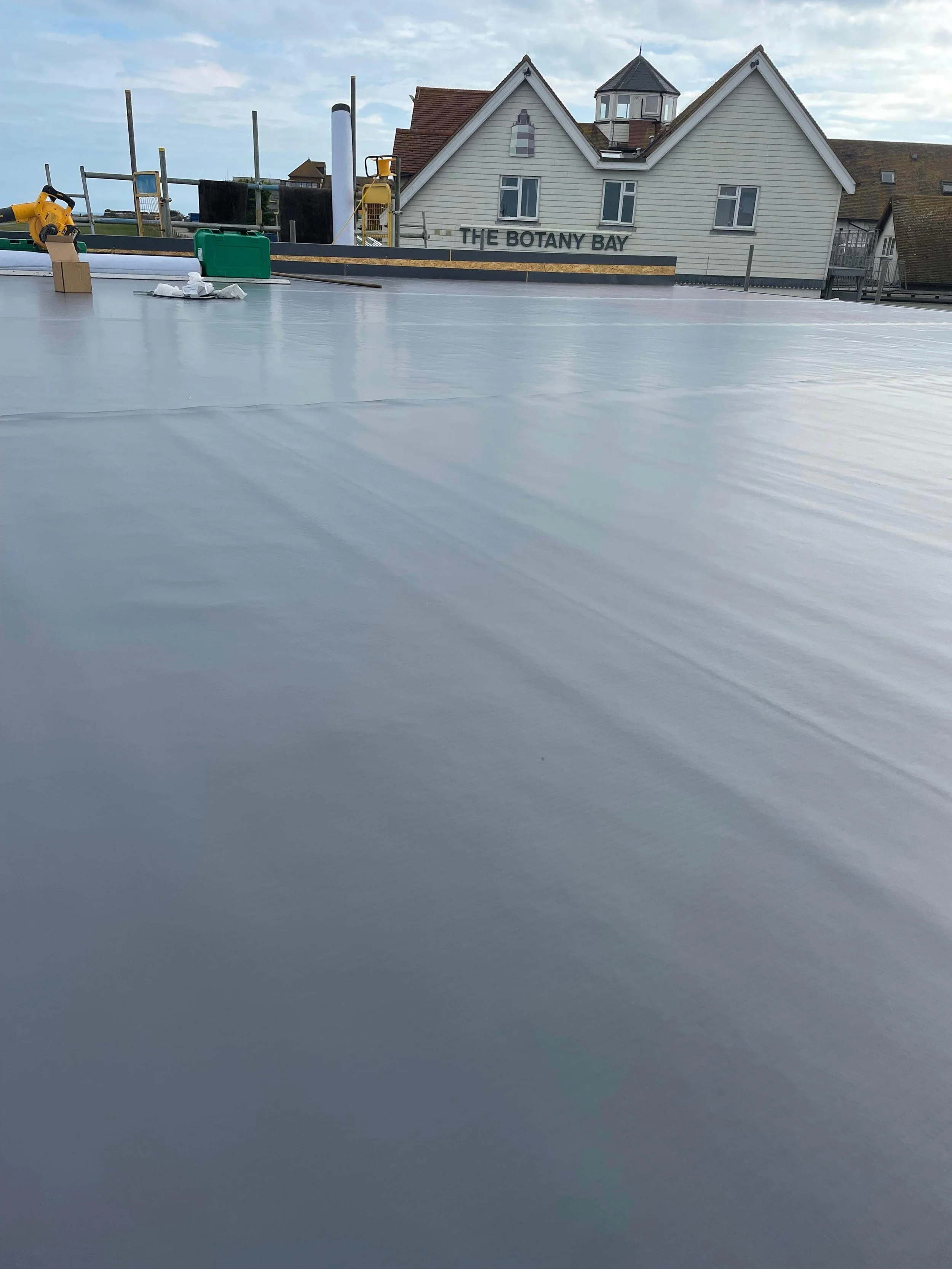 View of a flat roof with a smooth, gray surface and various tools and equipment near the edge, with house buildings and a cloudy sky in the background.