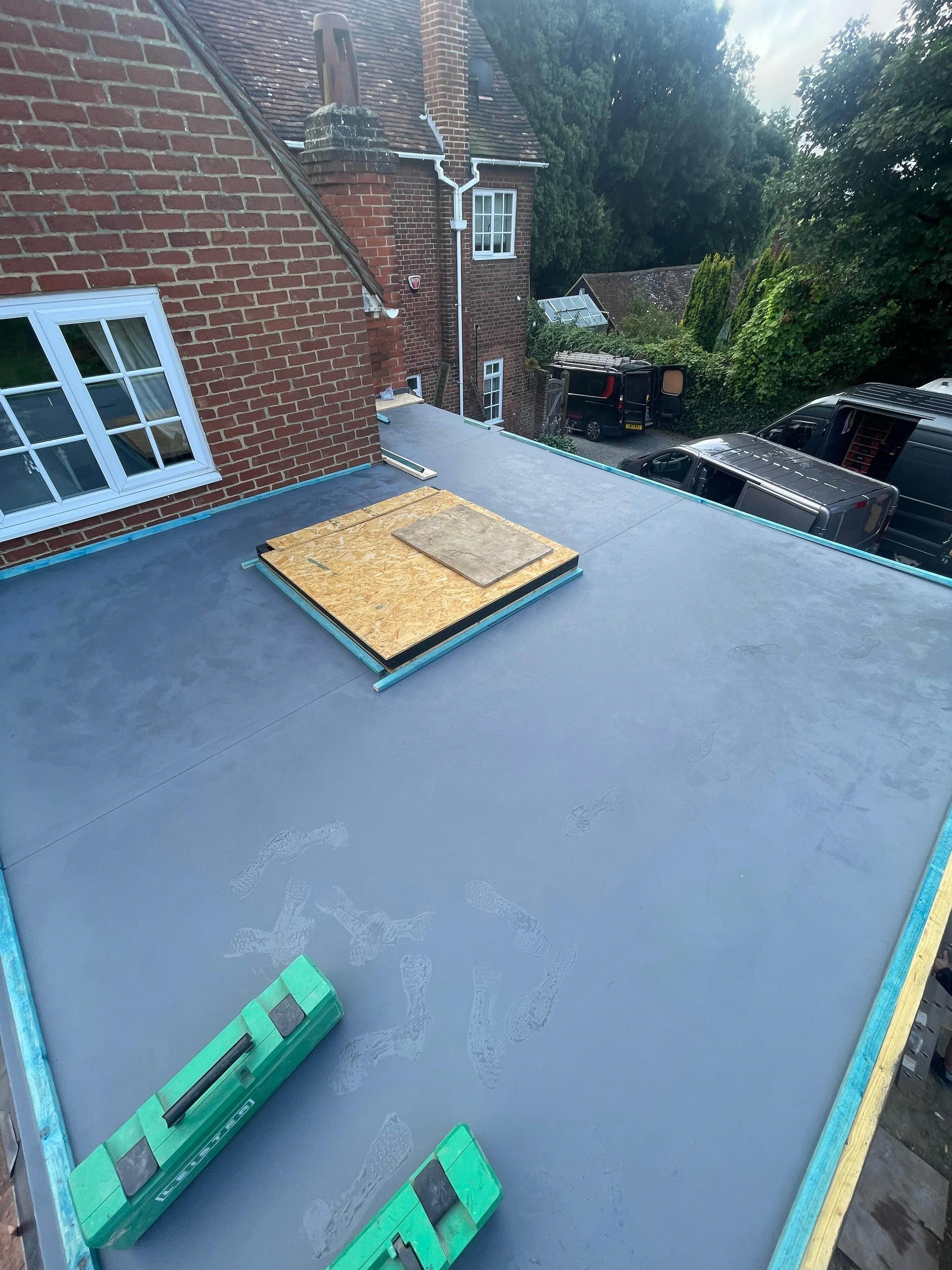 Flat roof under construction with a new waterproofing layer, construction tools, and wooden boards, overlooking brick houses, trees, and parked vehicles.