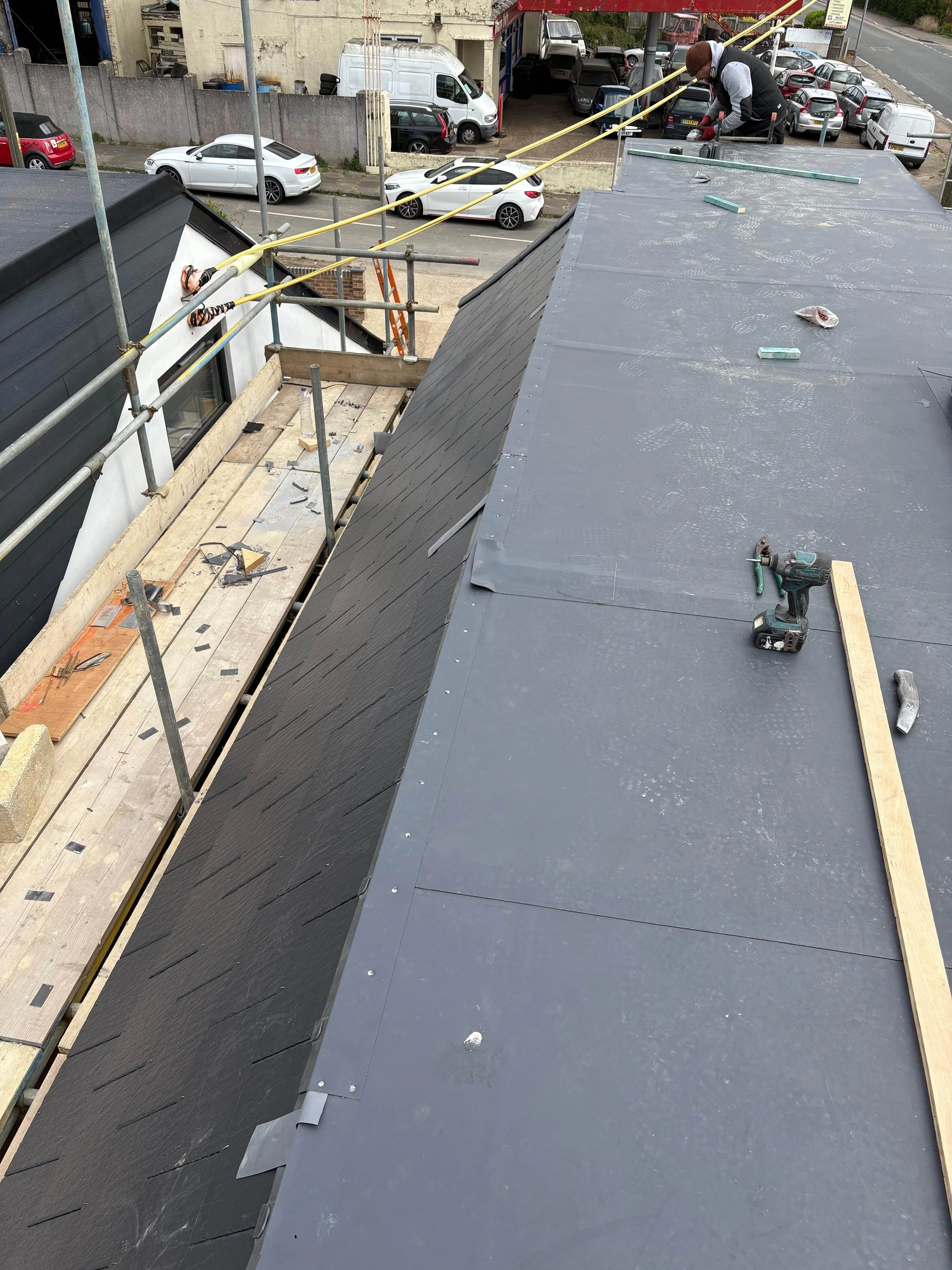 Construction worker on rooftop installing or repairing a metal roof with tools and supplies scattered around.