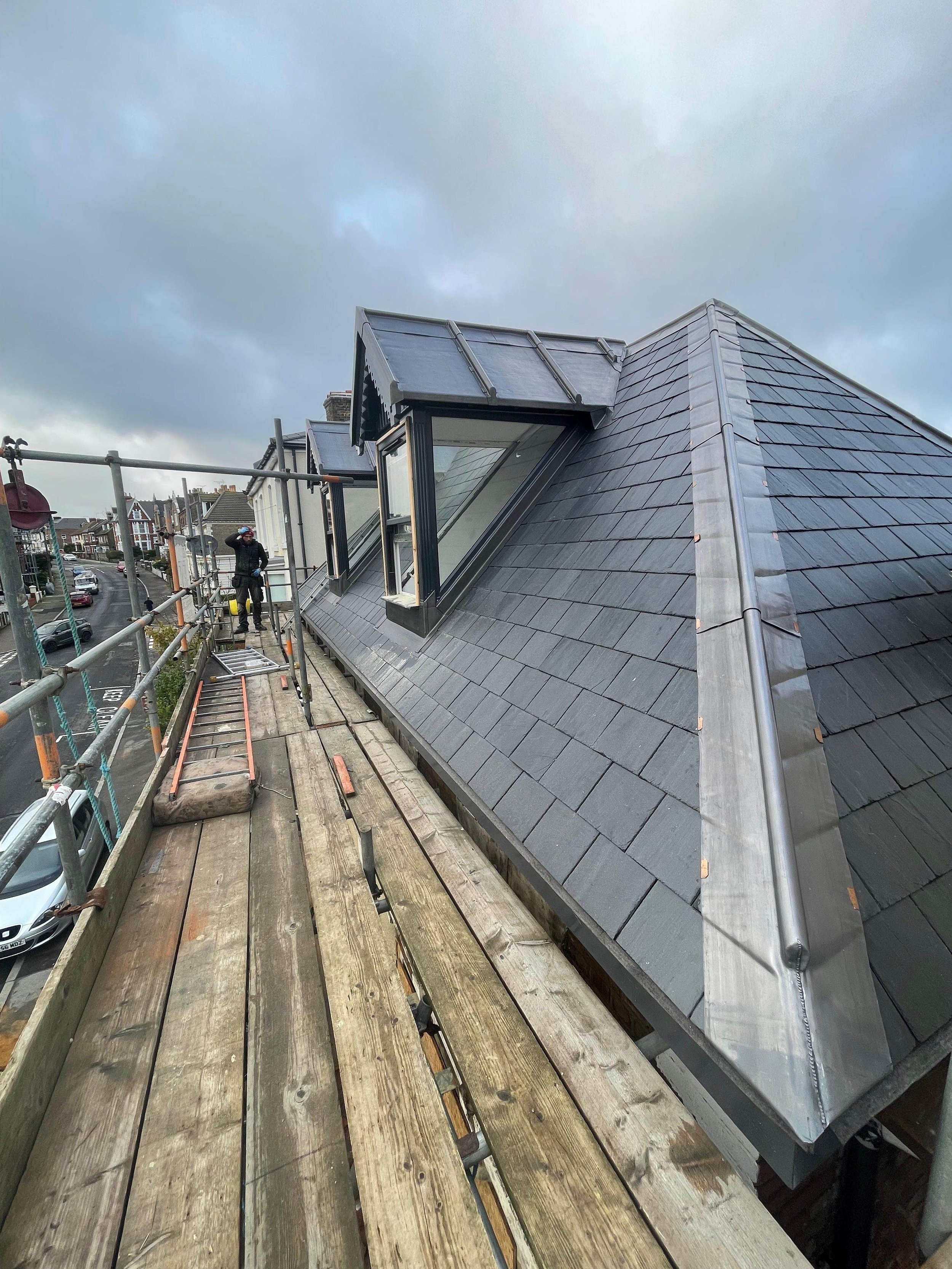 Construction worker standing on scaffolding next to a sloped roof with gray shingles and large windows, overlooking a residential street on an overcast day.