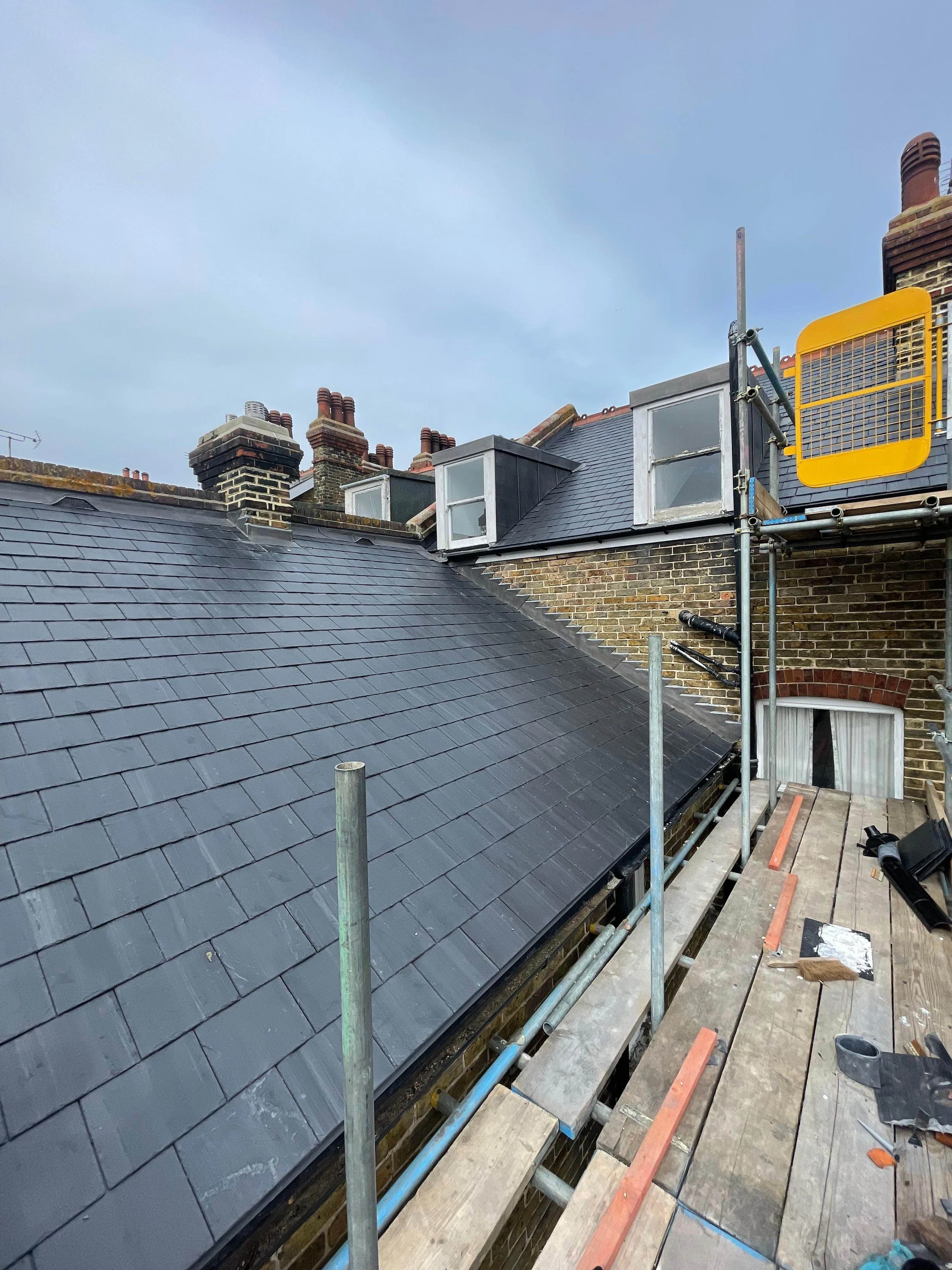 Roof renovation site with scaffolding and new black slate roof tiles on a brick building. Construction tools and materials are scattered on the wooden scaffolding platform.