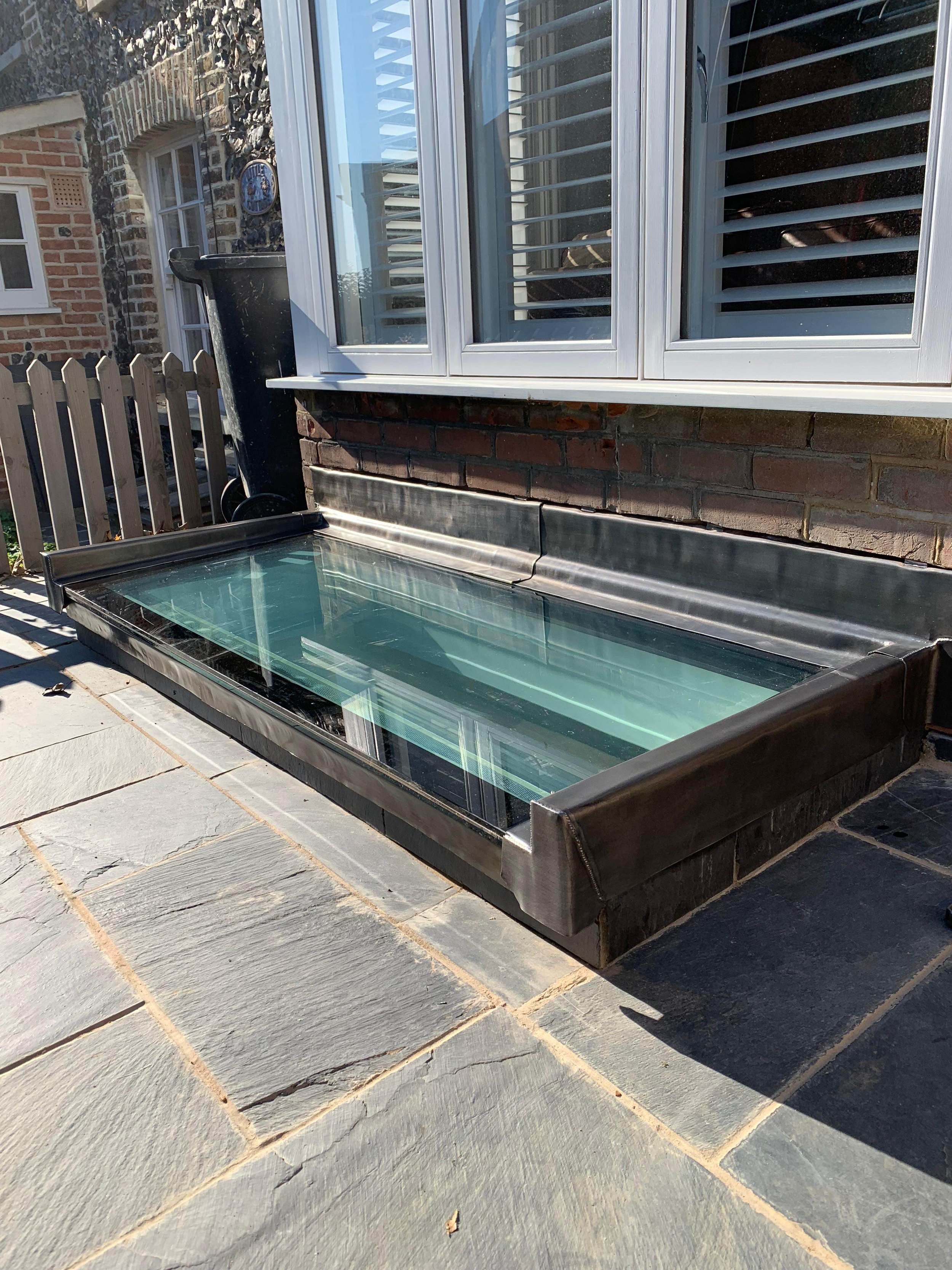 A rectangular glass skylight with a metal frame installed on a stone patio next to a brick house with white-framed windows.