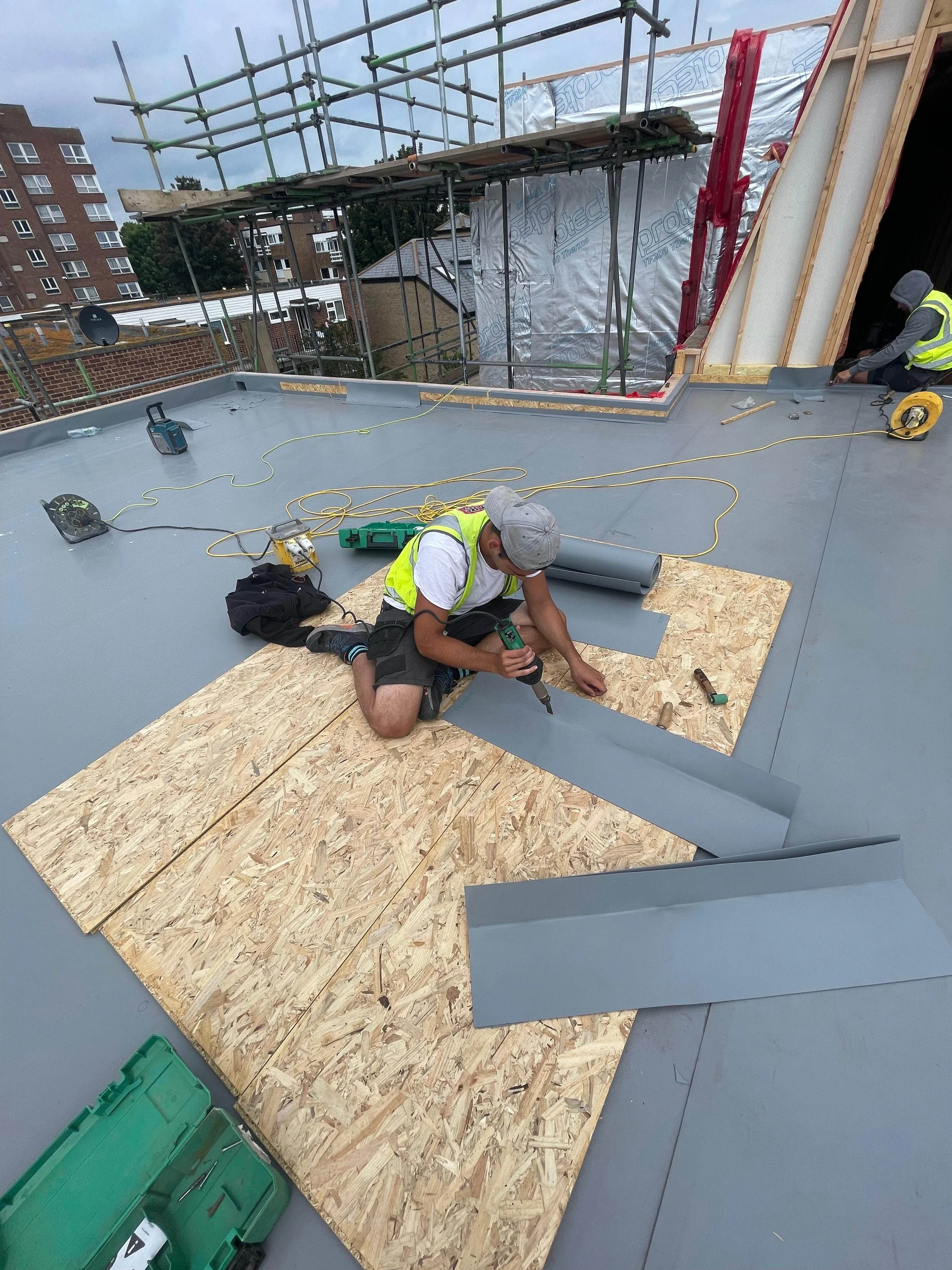 Construction workers installing roofing materials on a flat roof. One worker is kneeling and working with gray metal sheets, while another is in the background near a partially covered wall. Scaffolding is visible around the perimeter, and tools and 