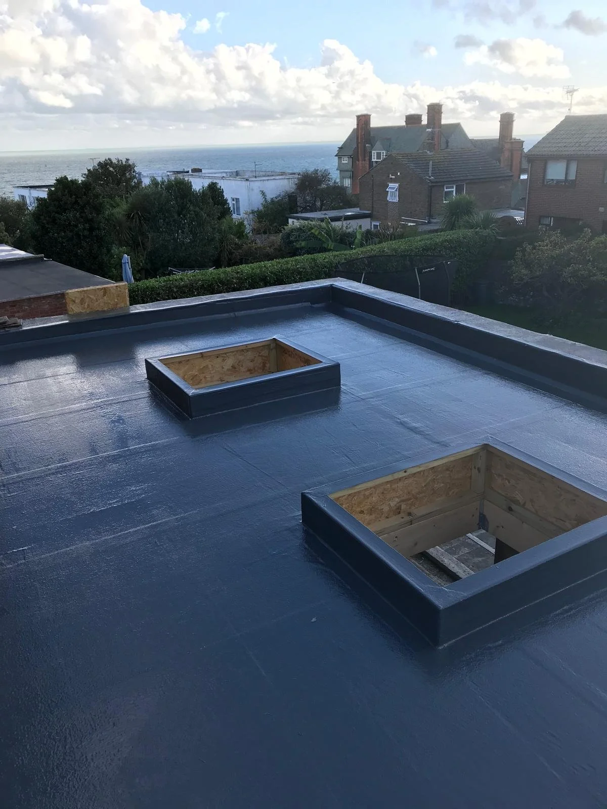 View from a flat rooftop with two square skylight openings, over the ocean and residential houses in the background, under a partly cloudy sky.