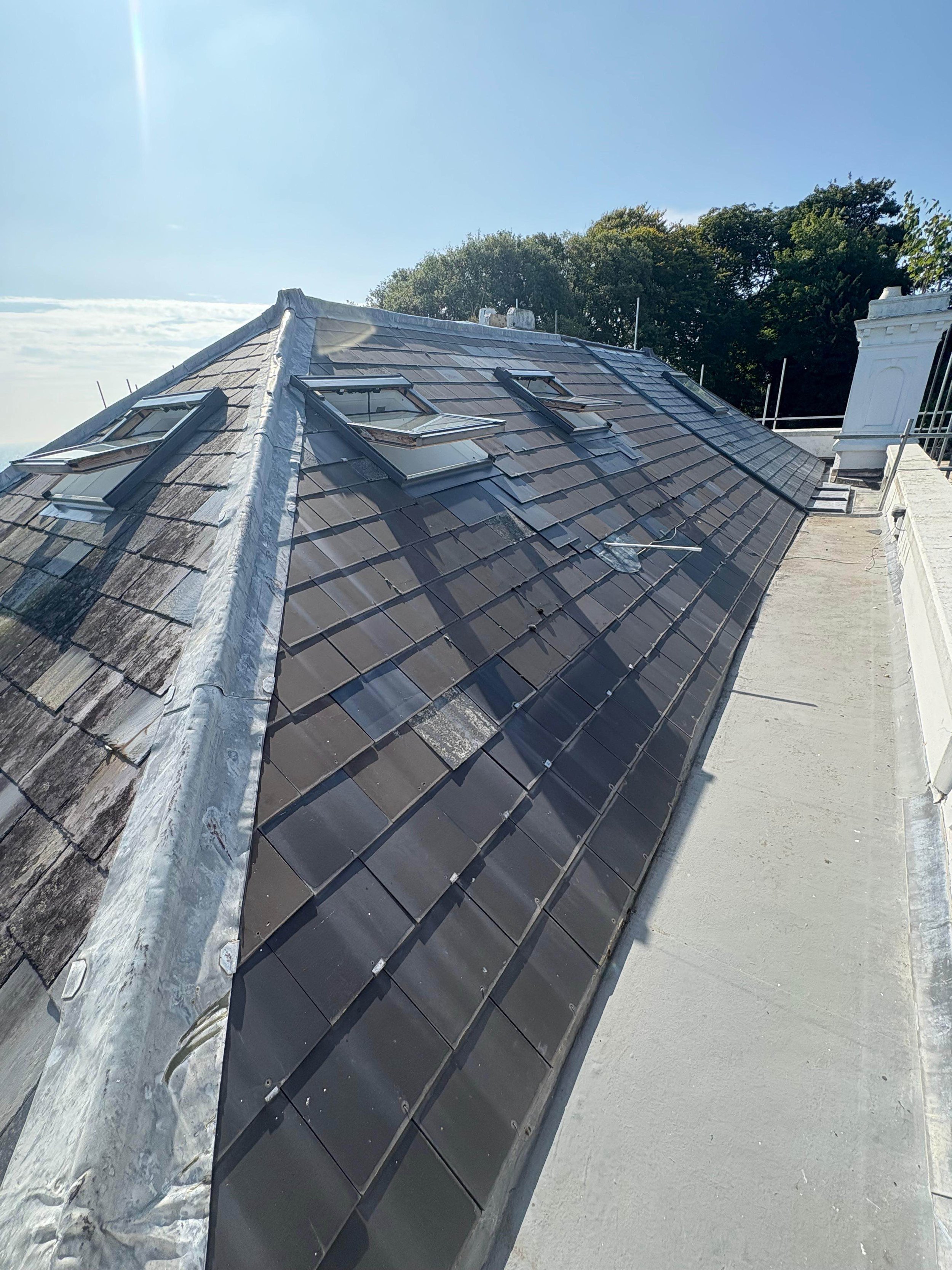 View of a sloped roof with black tiles, four skylights, and a chimney, with trees in the background and a bright blue sky.