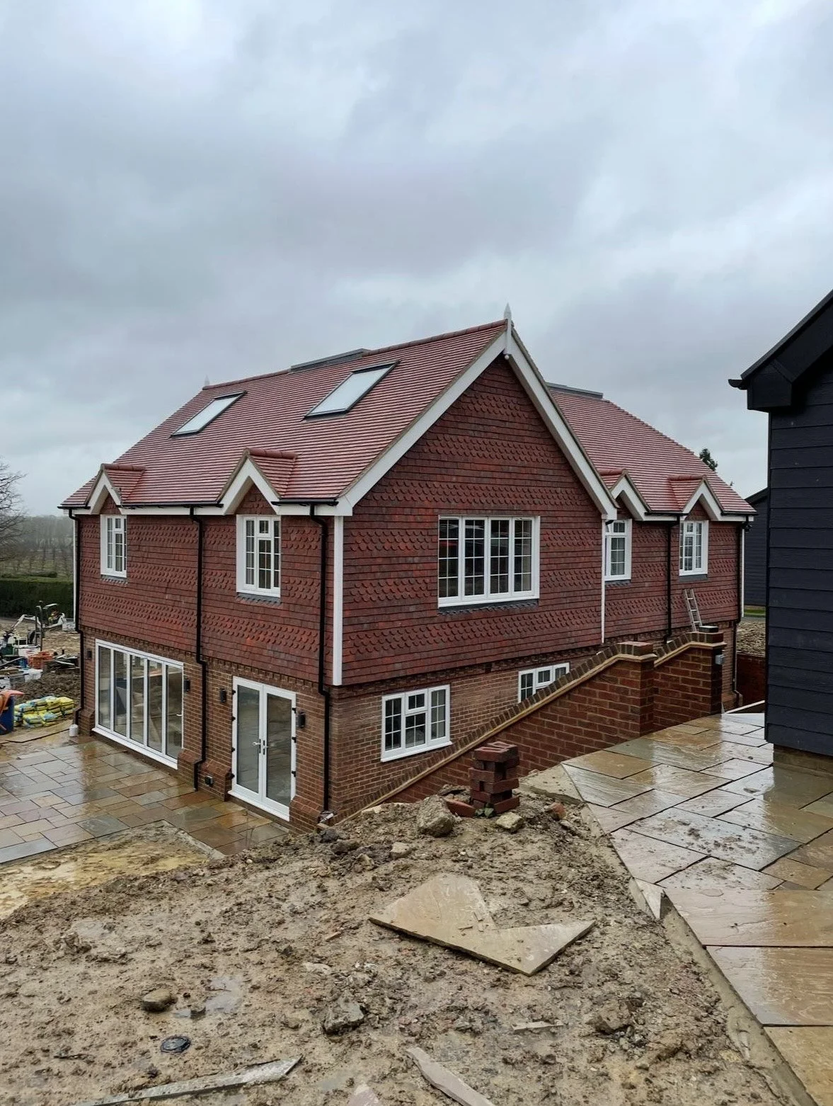 A two-story house under construction with red brick and brown shingle siding, white-framed windows, a sloped roof with skylights, and a tiled patio in the backyard on a cloudy day.