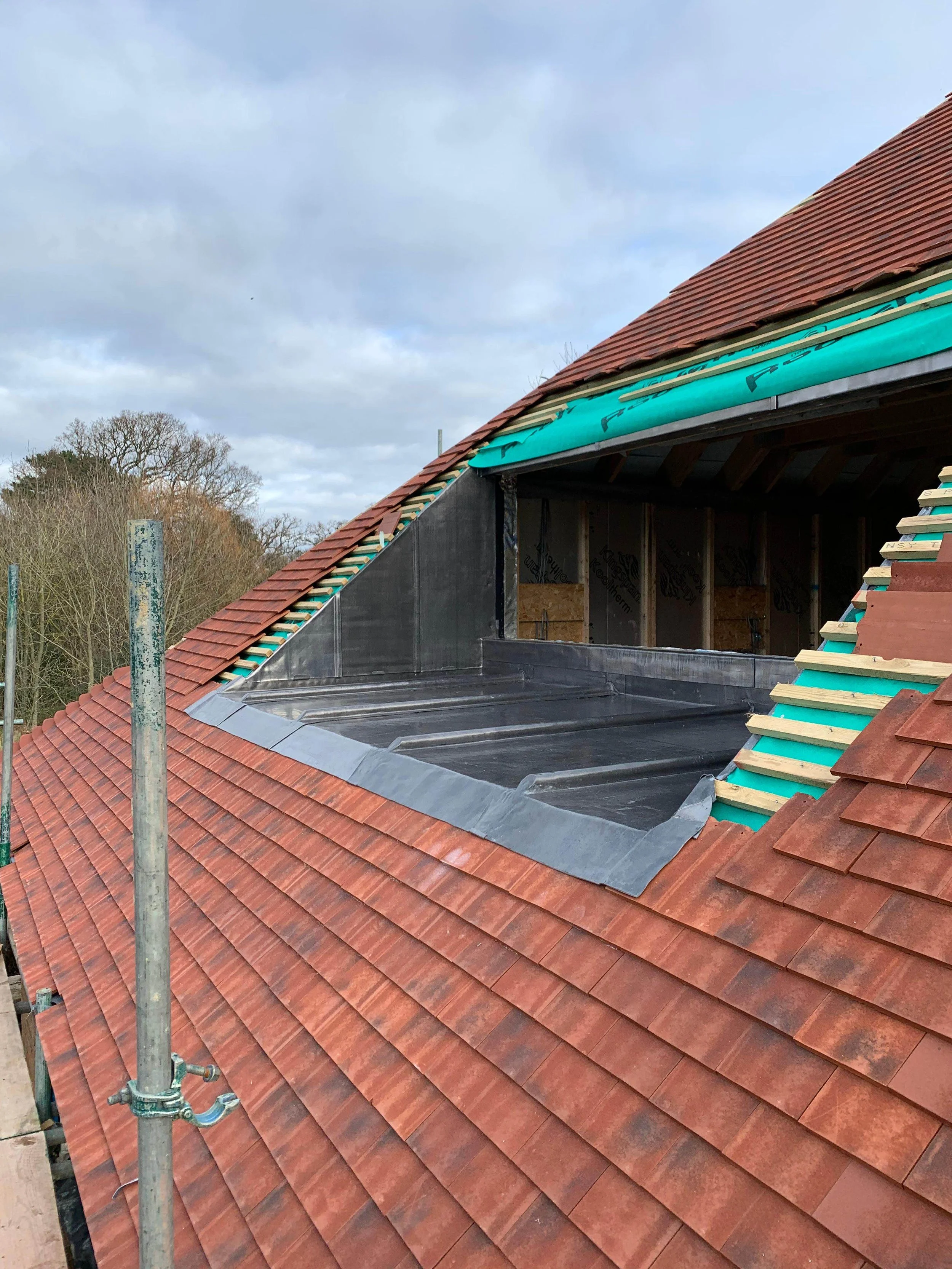 A house under construction with a red tile roof, where a section of the roof is being repaired or replaced. The roof is partially open, showing a new black waterproof membrane and wooden framing. There is scaffolding on the left side of the image, an