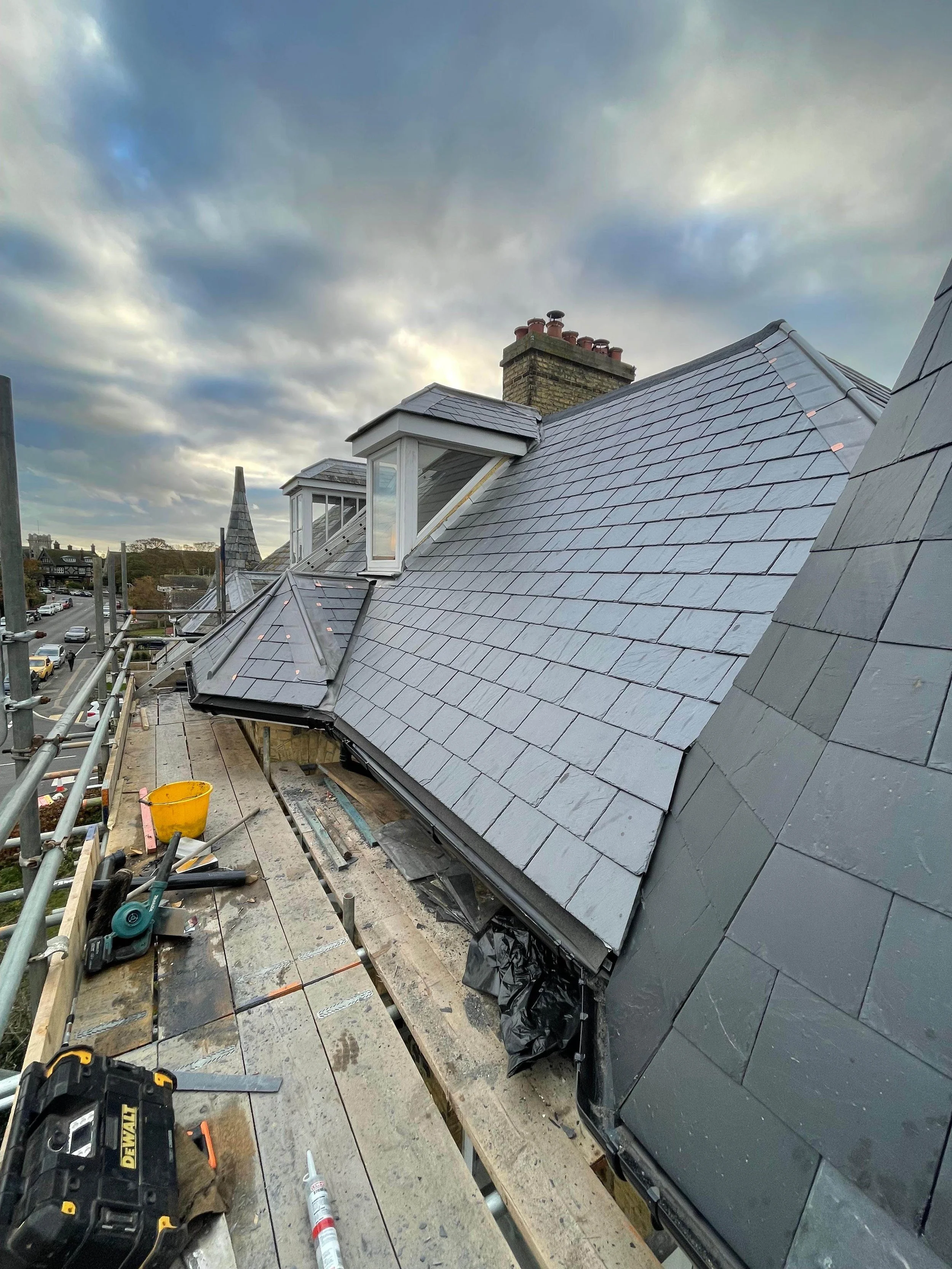 Construction or repair work on a rooftop with slate shingles, tools, and construction materials resting on scaffolding, under cloudy sky.